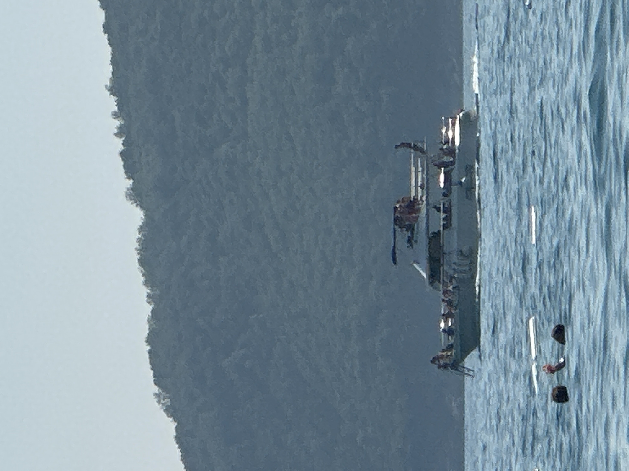 A yacht with several people aboard navigates choppy, blue water with a lush, green hillside in the background. In the foreground, three figures, two dark and one with a red object, are partially submerged in the water. The yacht appears to be moving towards the right side of the frame.