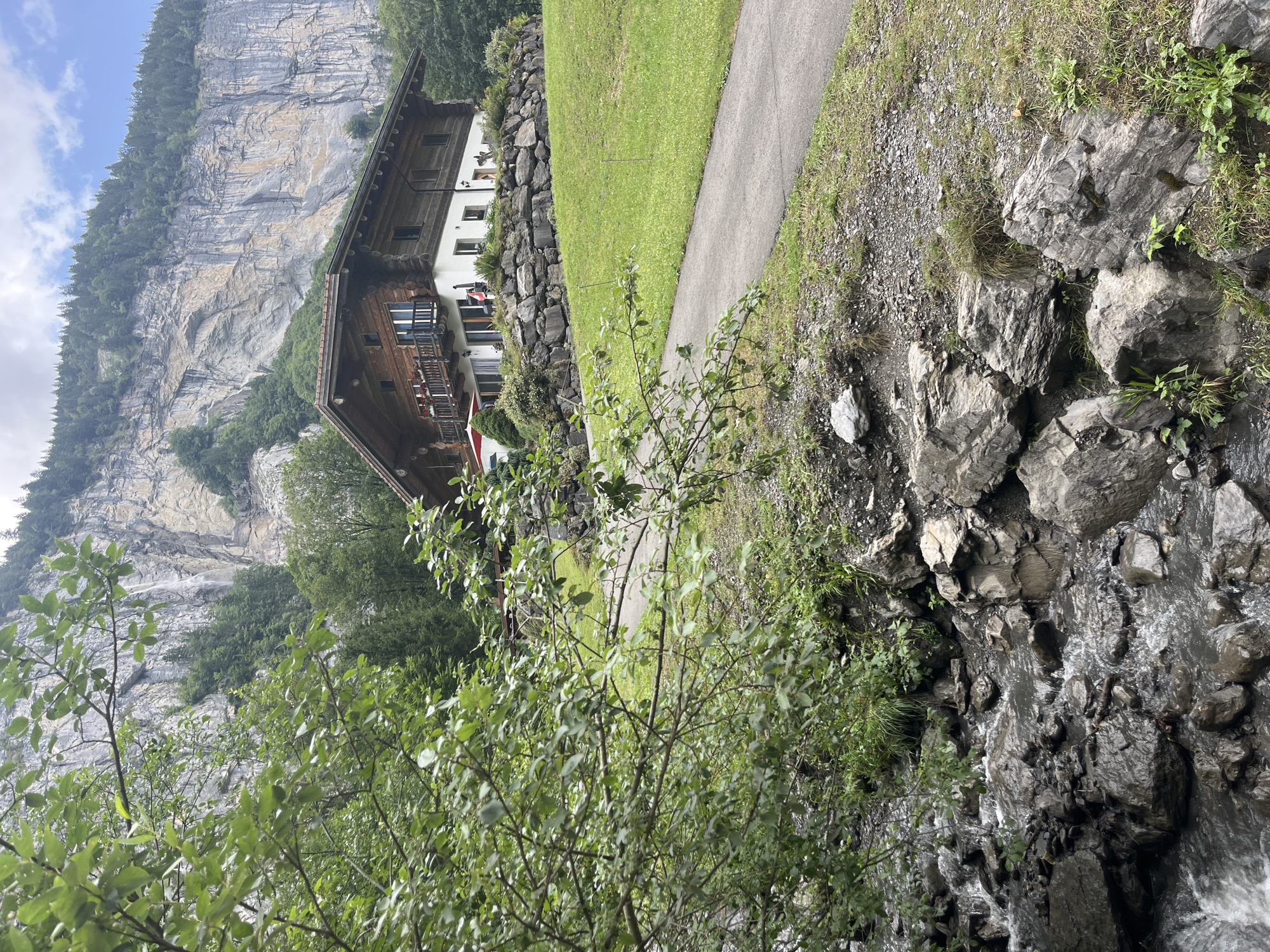 The image depicts a picturesque natural scene in Lauterbrunnen, Switzerland, characterized by lush greenery and dramatic geological formations. In the foreground, a rocky stream flows over dark, wet stones, with branches of green foliage extending from the left side of the frame, partially obscuring the view. A paved pathway winds its way through a manicured green lawn, leading towards a traditional Swiss chalet. The chalet, constructed primarily of wood with white stucco accents, sits on a terraced hillside. A stone wall, constructed from large, irregular rocks, supports the lawn in front of the chalet. Above the chalet, a towering, sheer cliff face rises majestically, its striated surface a testament to immense geological time. A thin waterfall cascades down the cliff face in the distance, barely visible through the trees. The sky above is partly cloudy, with patches of blue peeking through. No people are visible in the scene, and the overall atmosphere is one of serene, unspoiled natural beauty.