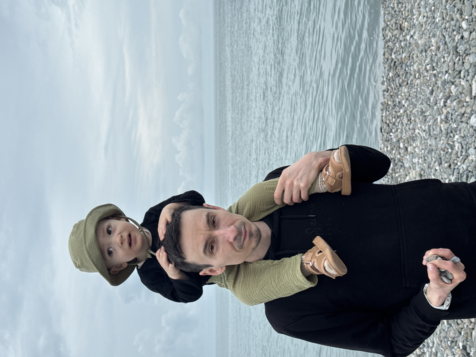 A medium-level, horizontally-oriented shot shows a man carrying a toddler on his shoulders. The man, fair-skinned with short dark hair and a light goatee, looks at the camera with a slight smile. He wears a black hooded sweatshirt. The toddler, fair-skinned, with dark hair and wearing a dark shirt and a green bucket hat, also looks at the camera. The child's legs are around the man's neck. The man's hands are visible, holding pebbles. The background shows a stony beach, the sea, and a cloudy sky. The scene is likely set in a coastal area, possibly Sochi, Russia, given the beach and sea context.