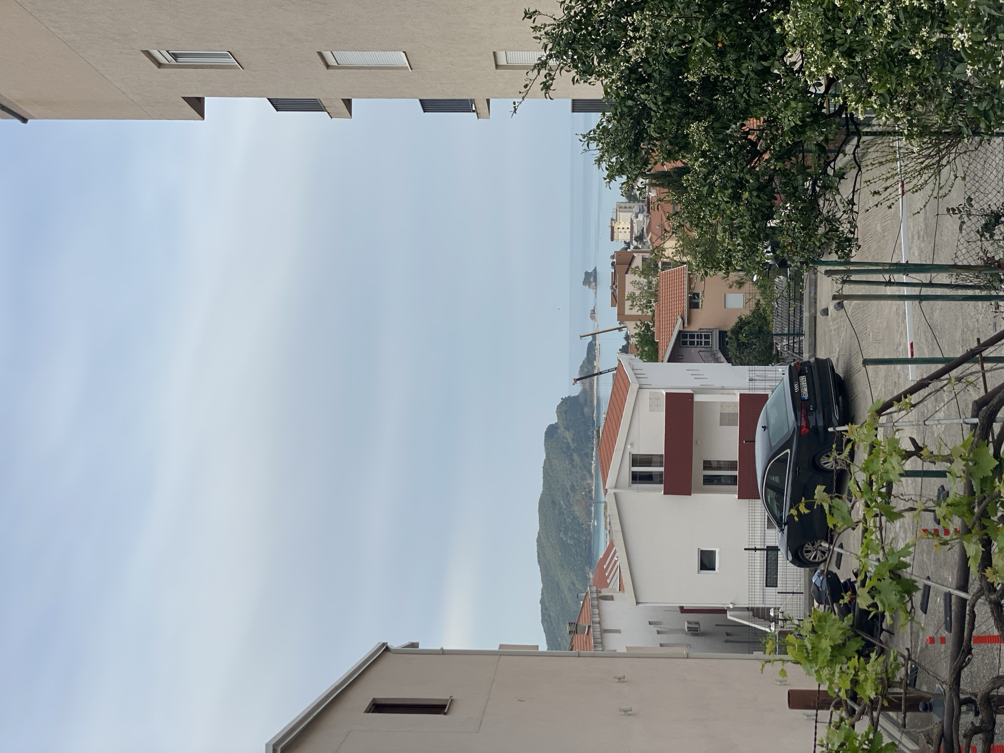 The image shows a view from between buildings in Budva, Montenegro, looking toward the sea. The sky is overcast. Buildings with red-tiled roofs are visible. A black Audi car (license plate: BD-BG524) is parked next to a white building with red-brown accents. A fence and a vine with green leaves are visible in the foreground. Mountains and the Adriatic Sea are visible in the distance.