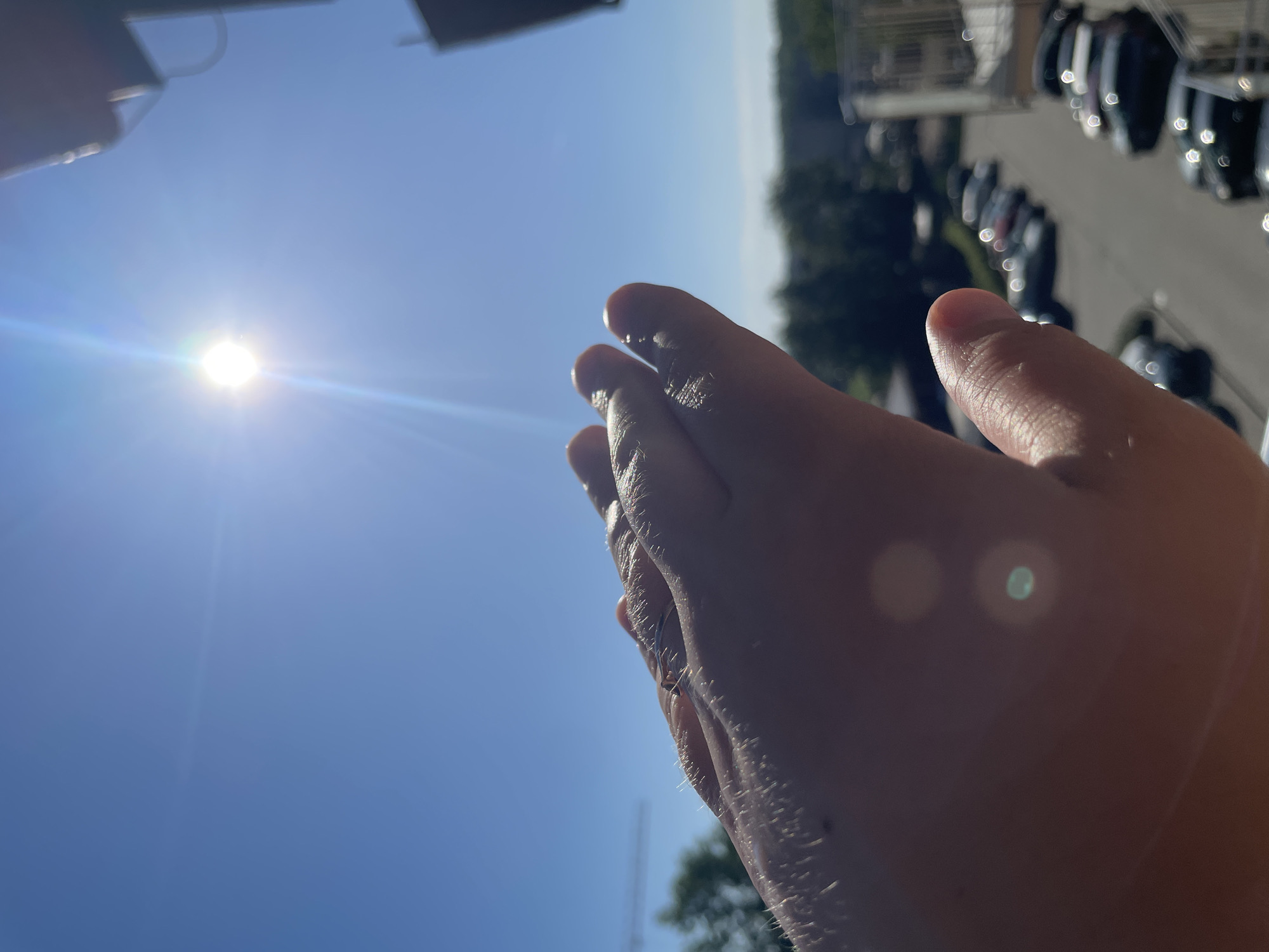 The image depicts a person's hand with outstretched fingers positioned against a bright blue sky with the sun prominently visible. A parking lot with several parked cars and some greenery are visible in the background. The location appears to be an urban environment, though the city is unknown. The hand is the main subject, positioned to partially obscure the sun. The sunlight creates a glare and strong contrast.