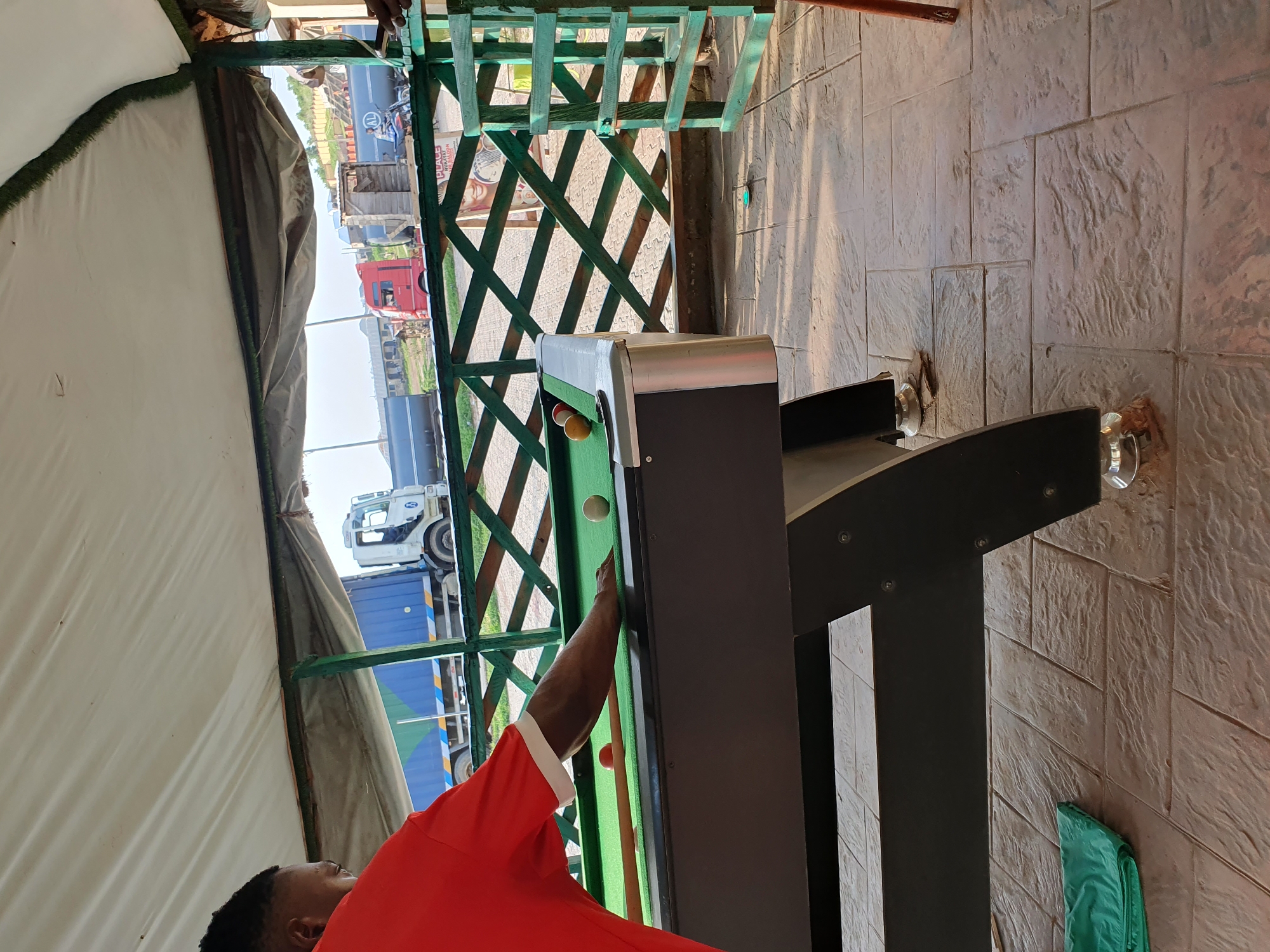 A person, wearing a red shirt with white trim, plays billiards on a green felt pool table. The table has a dark gray frame and a metallic edge. Two billiard balls, one white and one orange, are visible on the table. A billiard cue stick is also present. The scene appears to be outdoors, possibly under a shaded canopy or structure with a white fabric roof. In the background, several large trucks are visible, some of them appear to be in motion or parked. One truck is white with a blue container, and another is red. The ground is paved with textured tiles that resemble stone. The time of day seems to be daytime, with shadows cast on the ground, suggesting sunlight. The overall setting suggests a casual outdoor recreational area with a view of a road or transport hub. There is no visible text in the image.