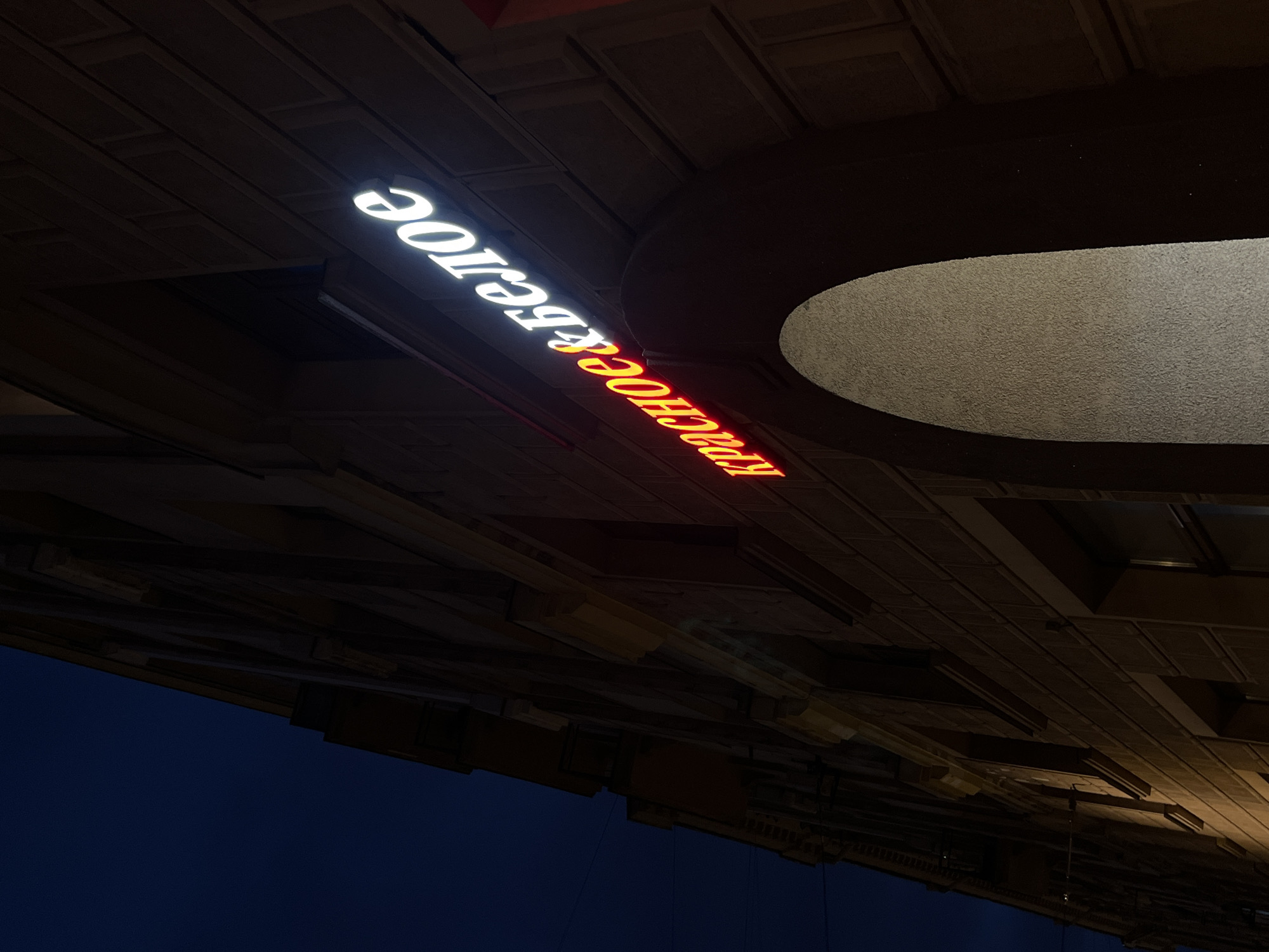 The image shows a neon sign on the side of a building in Saint Petersburg, Russia. The sign, in Cyrillic script, reads "Красное&Белое" which translates to "Red&White". The letters are illuminated with a red and white glow, suggesting it is a store or establishment. The building appears to be made of brick and has architectural details common in Saint Petersburg. The sky is dark blue, indicating it is nighttime. The sign is positioned above an arched entrance to the building. There are no people visible in the frame.