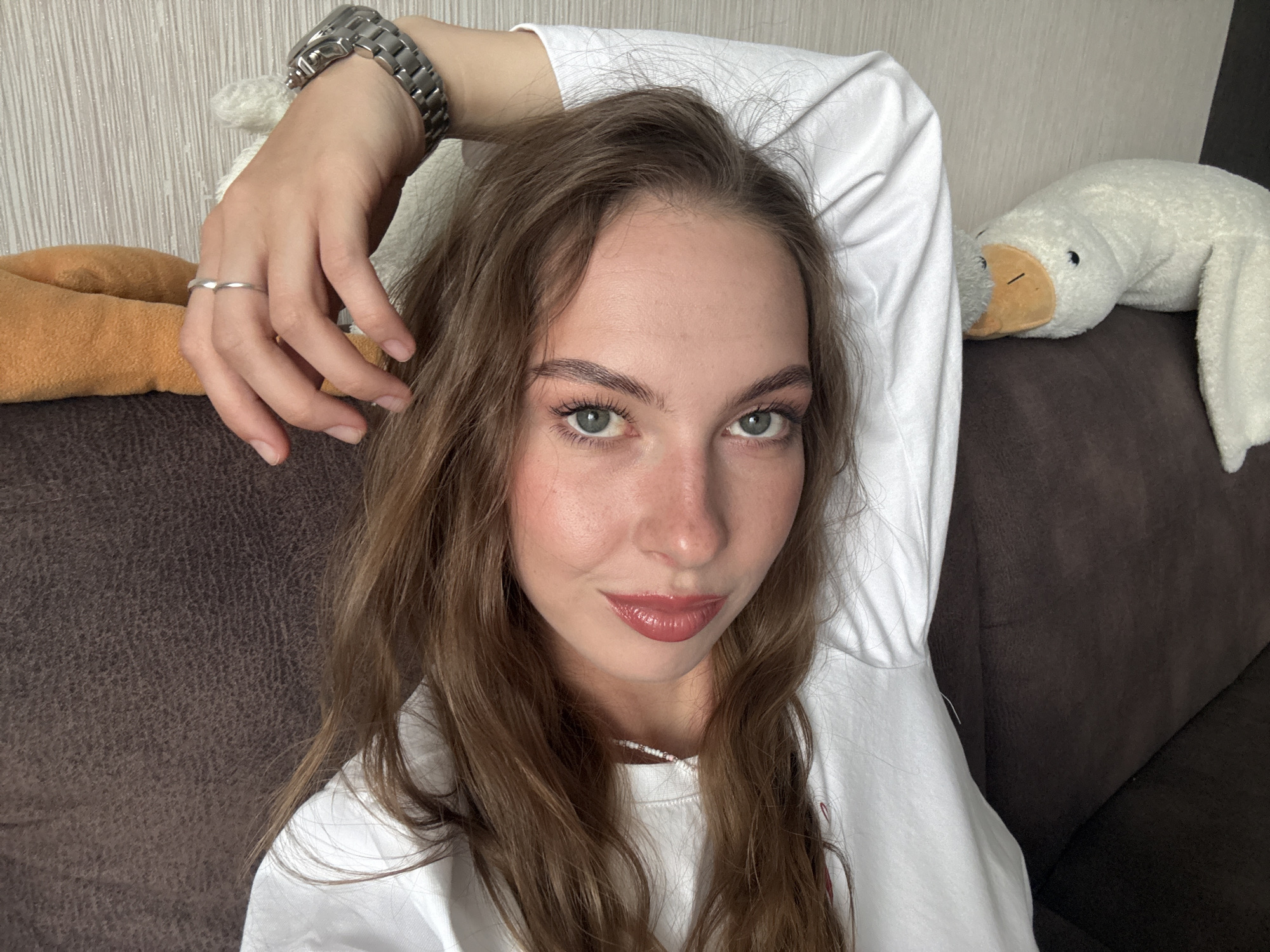 A front-facing, waist-up selfie depicts a young adult female reclining on a dark brown, textured sofa in an indoor setting, likely a residence in the city of Perm, Russia. The subject has long, wavy, light brown hair parted on the left side, framing her face and extending over her shoulders. She has light green eyes with visible mascara, well-defined arched eyebrows, a straight nose with a subtle reddish hue on the bridge and tip, and full lips coated in a reddish-pink glossy lipstick, displaying a faint smile. Her complexion is fair with some faint freckles on her nose and cheeks. She wears a white long-sleeved crew-neck sweatshirt or t-shirt, a silver-toned metal link watch on her right wrist, and a simple silver-toned band ring on her left ring finger. A white beaded necklace with interspersed darker beads is visible around her neck. Her right arm is raised and bent, with her hand positioned near her hairline.
To the subject's left, a portion of an orange or tan soft object, possibly a blanket or plush toy, is visible on the sofa. To her right, a white plush toy resembling a platypus, with an orange beak and small black eyes, rests on the sofa. The background consists of a light-colored wall featuring a subtle vertical textured wallpaper pattern. The lighting is soft and even, suggesting an interior daytime environment.