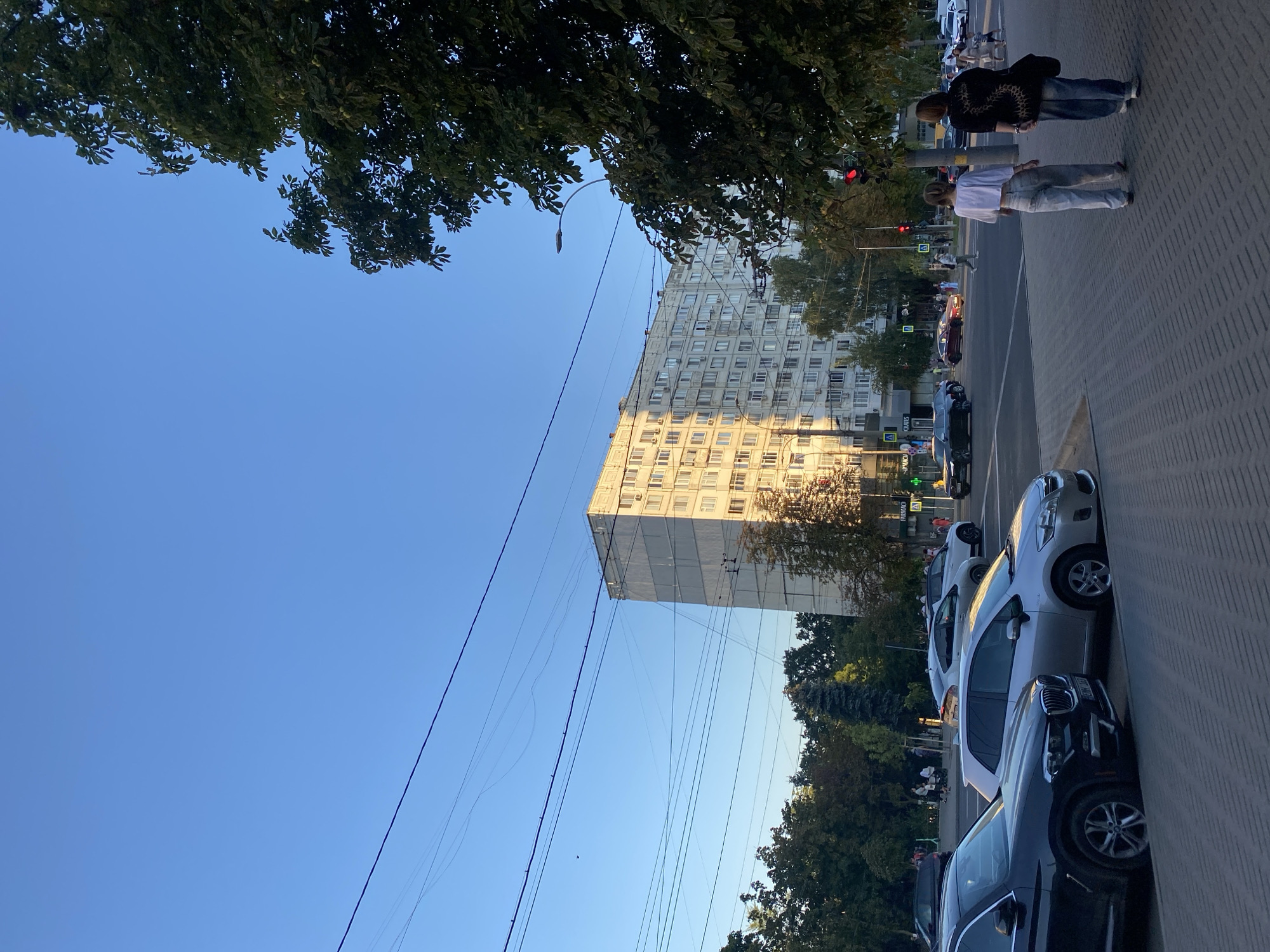 The image captures a street scene in Chișinău, Moldova, during daylight hours with a clear blue sky. The foreground features a paved sidewalk with two individuals walking away from the camera. The person on the left wears a white short-sleeved top and light-colored pants, while the person on the right is dressed in a black patterned top and dark pants. To the left of the sidewalk, several parked cars are visible, including a black SUV and a silver hatchback.
Beyond the sidewalk, a street runs perpendicular to the camera's view, with traffic and pedestrians. A large, multi-story apartment building with a light-colored facade dominates the background, its windows reflecting the sunlight. Trees line the street and surround the building, some displaying lush green foliage. Overhead, numerous power lines crisscross the sky. Traffic lights are visible, with one showing red. The overall impression is a typical urban environment in the late afternoon or early evening.