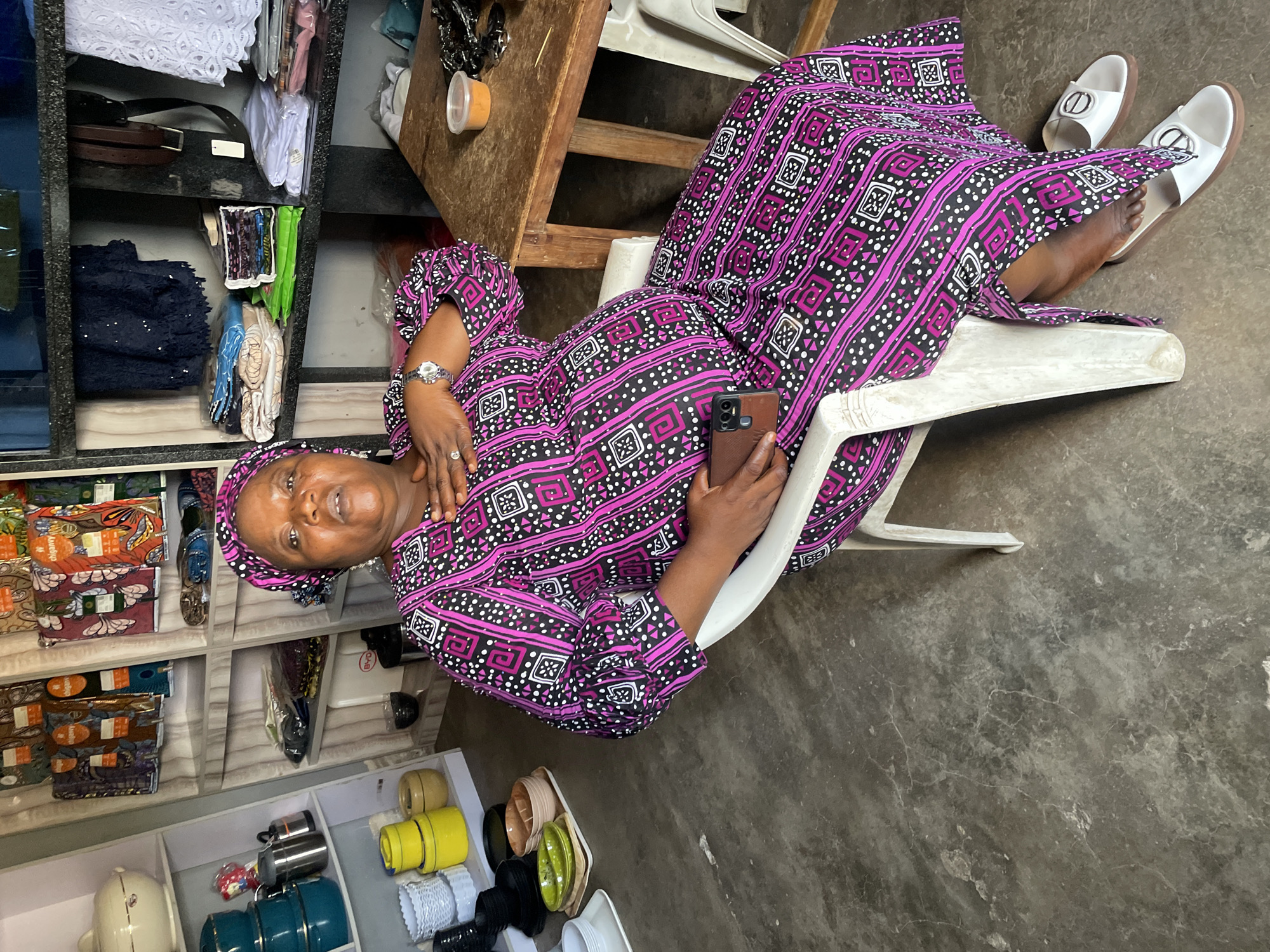 An adult woman in patterned clothing sits on a white plastic chair, holding a smartphone in her right hand and touching her neck with her left. She is wearing sandals and a wristwatch. The setting appears to be a retail space or shop. A shelf unit with various items, including fabric, belts, and containers, is behind her. A small wooden table with a container sits beside the chair. The location is potentially in Jos, Nigeria, considering the subject's clothing and general appearance, though there is no definitive evidence in the image. The floor is a grey concrete or tile.