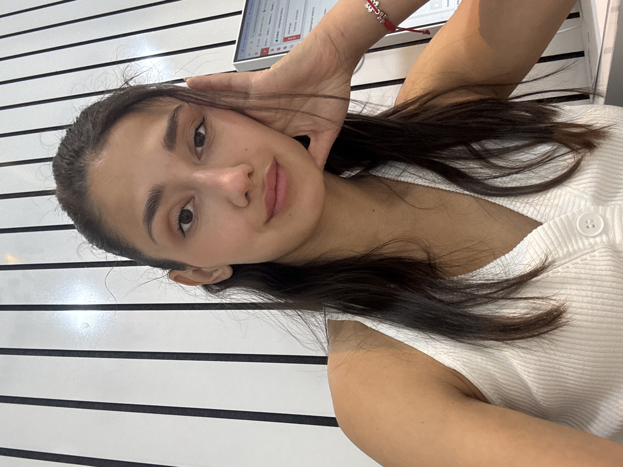 A young woman is pictured indoors against a backdrop of horizontal black and white stripes. She has dark brown hair and eyes and fair skin. She wears a white ribbed sleeveless top. Her left hand is raised near her face, resting on her cheek. Part of a tablet computer is visible in the background. It is not possible to determine if the photo was taken in Podgorica, Montenegro, without additional information.