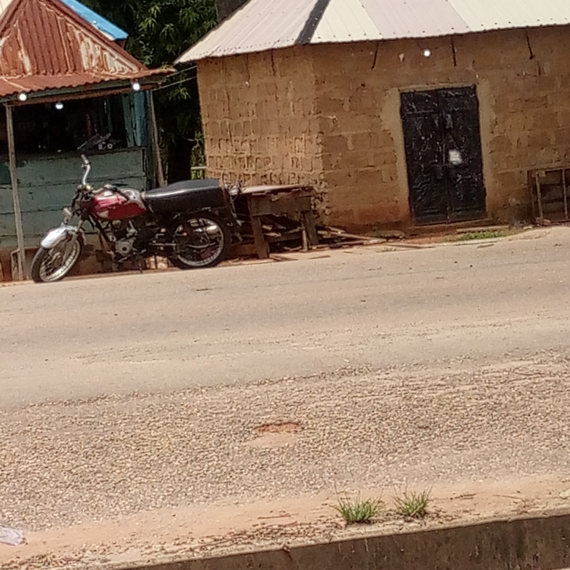 The image displays an outdoor scene in what appears to be a semi-urban or rural area, consistent with Mararaba, Nigeria. A red and black motorcycle, possibly adapted for transport with a rear carrier or small sidecar, is parked on the left side of a dirt and gravel road. The road surface is uneven, with loose stones and patches of earth. Along the immediate foreground, a concrete curb separates the road from a narrow strip of dirt with sparse green grass.
In the background, two modest buildings are visible. On the left, there is a structure with a heavily rusted, corrugated metal roof and an open-fronted design, suggesting a small stall or shop. Two small, circular light fixtures hang from its eaves. Behind this, lush green tree foliage is partially visible. To the right, a rectangular building constructed from unplastered mud bricks or blocks stands, topped with a lighter-colored corrugated metal roof. It features a dark, ornate metal door centrally positioned on its visible facade. Between the motorcycle and this brick building, a makeshift wooden table or stand is present, possibly used for commerce or as a workstation.
The scene is illuminated by natural daylight, suggesting it is mid-morning or afternoon under clear skies, with no visible signs of rain or heavy cloud cover. There are no people visible, and no specific activity or event is taking place, contributing to a quiet ambiance. No visible text is discernible in the image.
