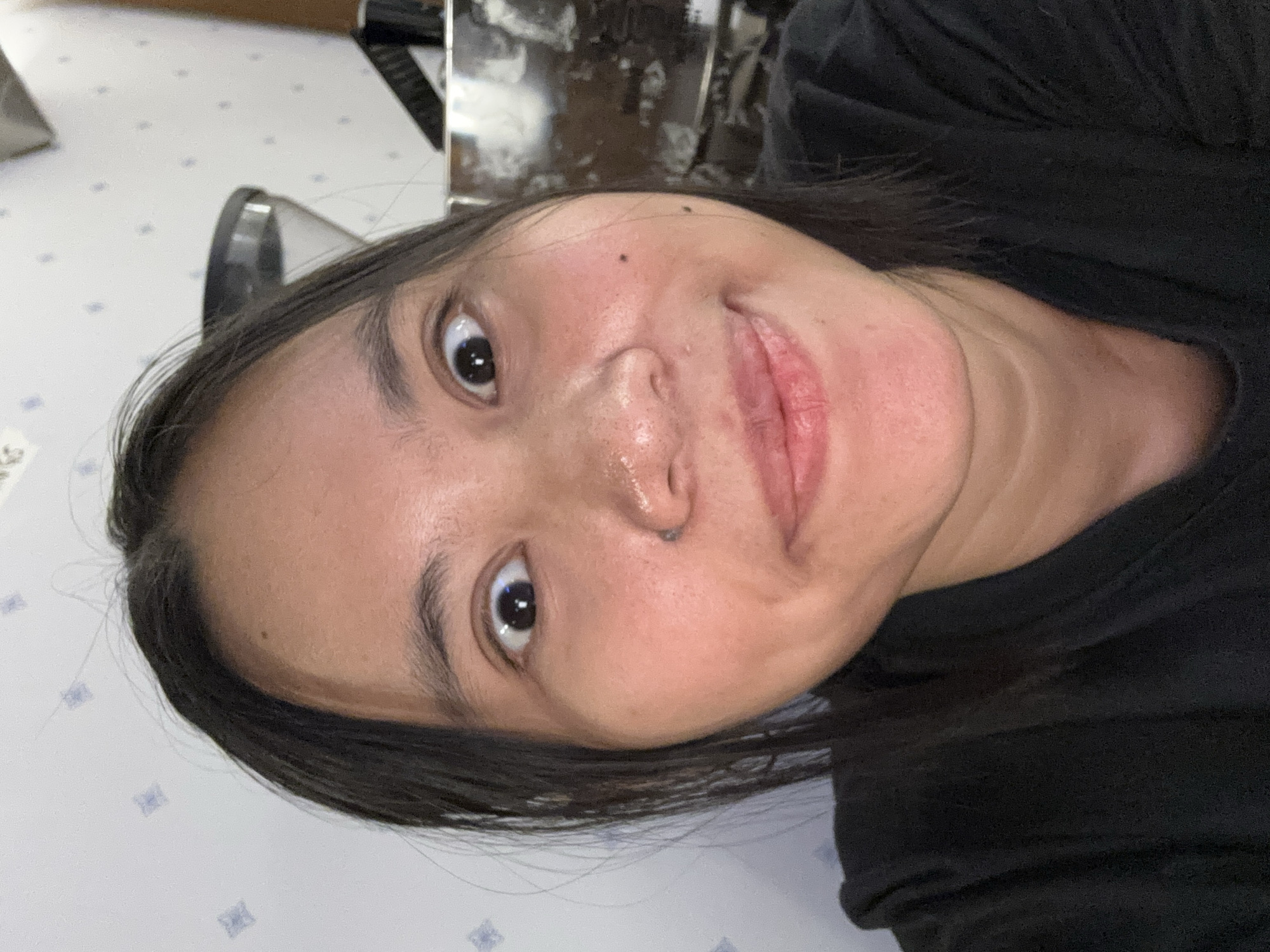 The image is a close-up selfie of a woman with dark hair, dark eyes, and fair skin. She is wearing a black t-shirt. Her expression is playful, with wide eyes and a slight smile. The background appears to be a domestic or indoor setting, possibly a home or cafe, with a patterned wall. There are no distinct landmarks or context to place the image specifically within South Jakarta, Indonesia.