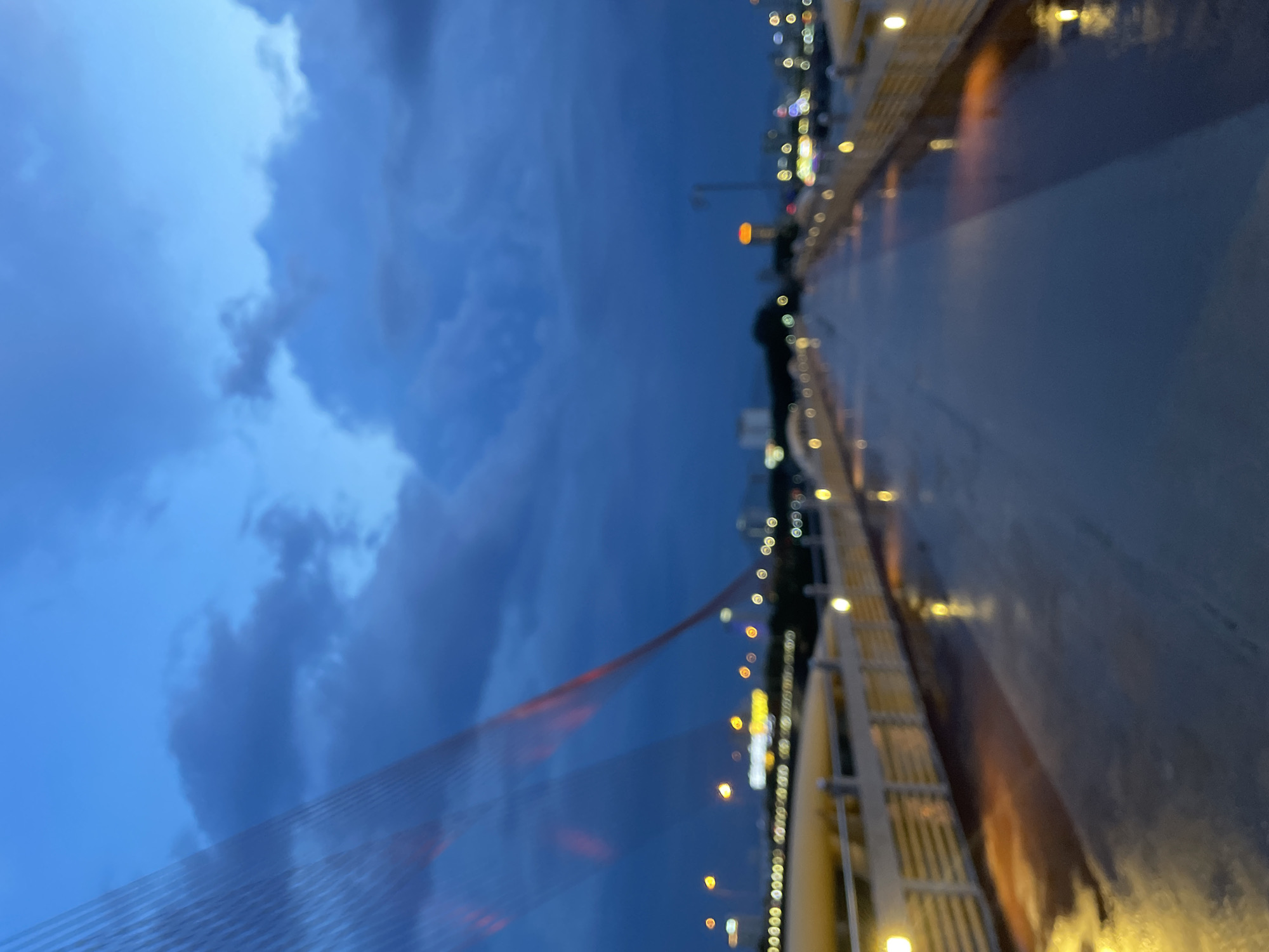 The image depicts a pedestrian walkway on the Dragon Bridge in Đà Nẵng, Vietnam, at dusk. The sky is a deep blue with dramatic, dark clouds. The bridge's iconic cable-stayed structure is visible in the upper left corner, with orange lights reflecting off the cables. The walkway itself is wet, indicating recent rain, and illuminated by warm yellow streetlights. In the distance, the city skyline is visible with blurred lights from buildings and vehicles. The overall impression is a moody and atmospheric scene of the city at twilight.