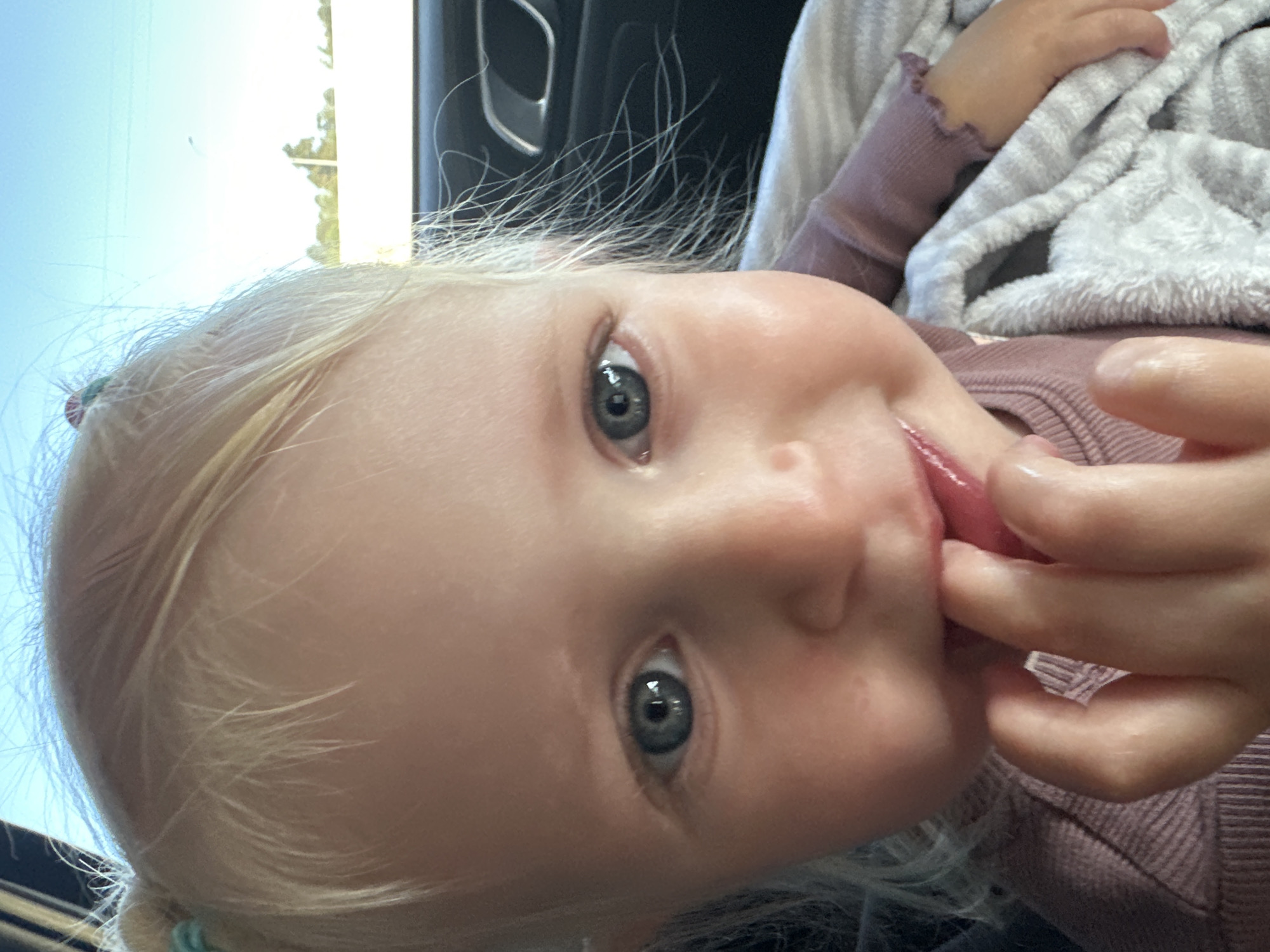 A close-up photograph shows a young child with light blonde hair and blue eyes. The child is wearing a long-sleeved purple shirt and is wrapped in a white, fuzzy blanket. The child is putting their fingers in their mouth. The background includes a car interior with a black handle visible. The scene appears to be inside a vehicle. No location information about Arsuf, Israel is apparent.
