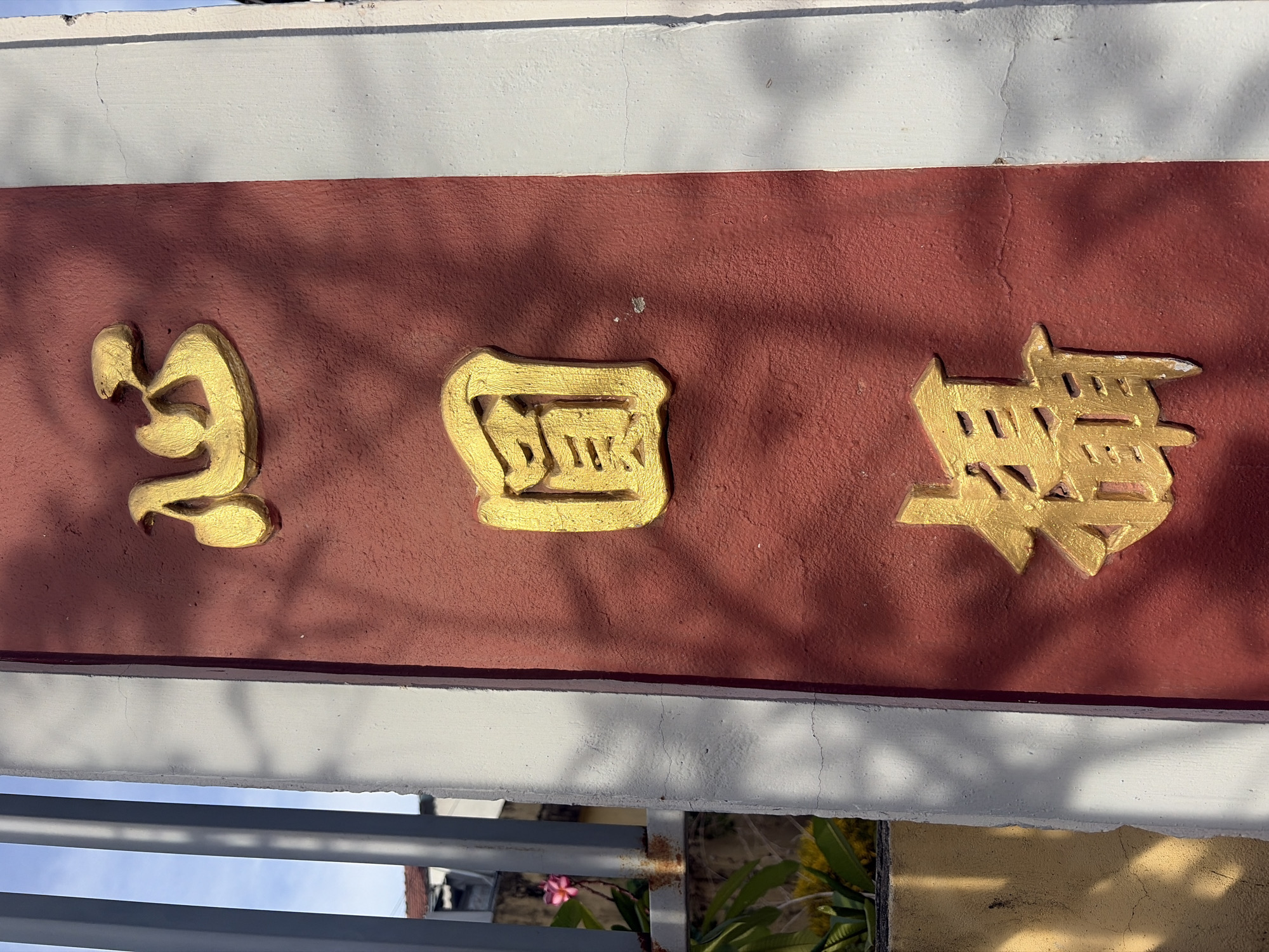The image shows a close-up of a red pillar with three golden Chinese characters vertically inscribed. The top character appears to be "心" (xīn), meaning heart or mind. The middle character is "圓" (yuán), meaning round or circle. The bottom character is "福" (fú), meaning fortune or blessing. The characters are embossed and appear to be made of metal, possibly brass or gold-plated. The pillar is textured and shows some minor cracking. Sunlight casts shadows across the surface of the pillar, highlighting the relief of the characters. To the left of the pillar, a white vertical trim is visible, and beyond that, a metal fence and some greenery, including a pink flower. The background suggests an outdoor setting with a bright sky. This pillar is located at the entrance of a temple or shrine in the city of Đà Nẵng, Vietnam.