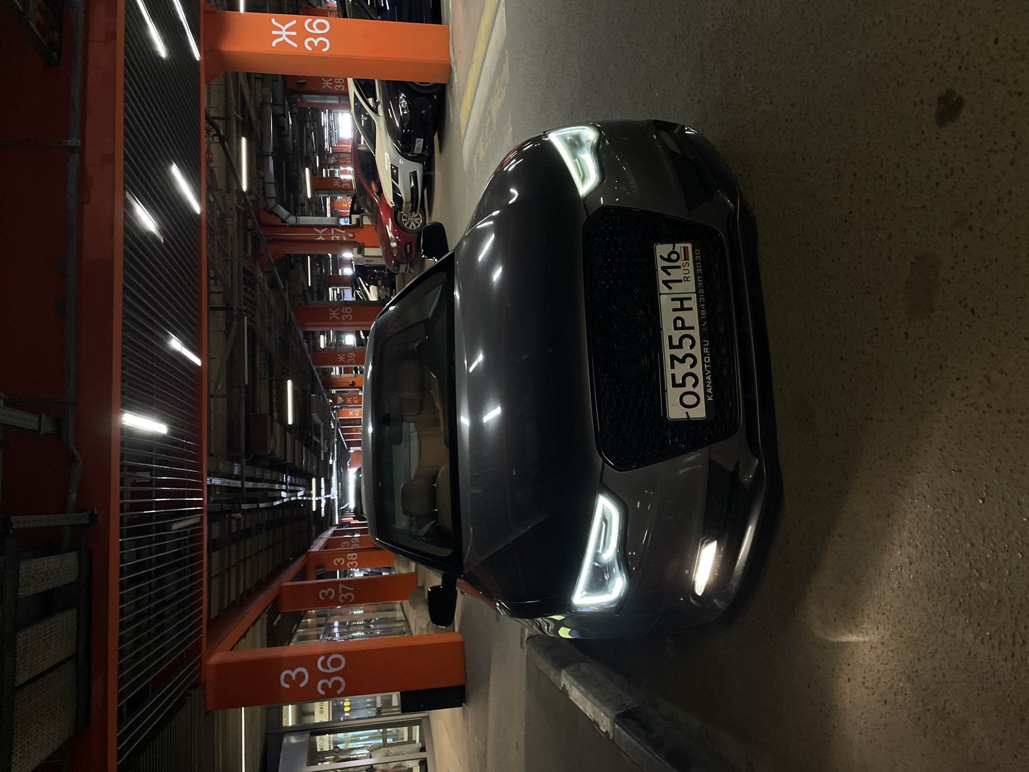 The image shows a gray car parked in an indoor parking garage. The car's license plate is visible and reads "0535PH 116" with "RUS" indicating Russia. The license plate also shows "KANAVTO.RU (843)230 30 30" written on it. The parking spot is marked with orange beams with the number 36 written on them, and other orange beams with numbers 37, 38, and 39 are visible in the background. The location is suggested to be Kazan, Russia, based on the license plate information. Several other vehicles can be seen parked further in the background. The overhead lighting in the garage is visible.