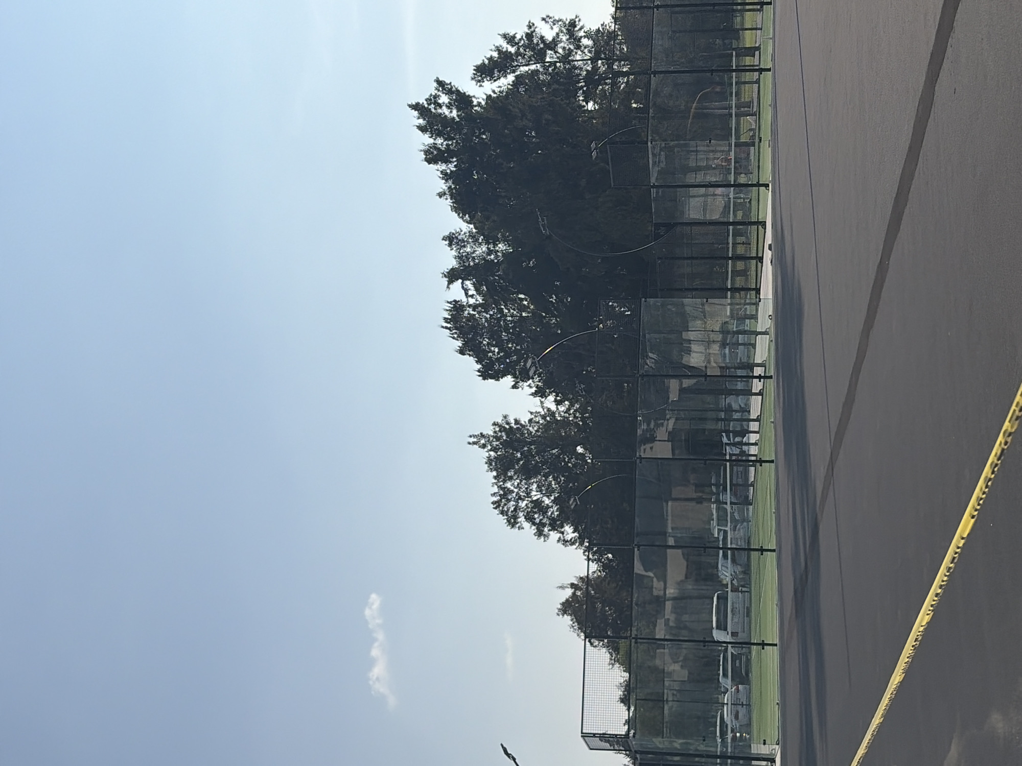 The image shows a row of paddle tennis courts in an outdoor setting, likely a recreational area. The courts are enclosed by wire mesh fencing, and trees are visible behind the courts. The playing surface is dark, possibly asphalt or a similar material, with painted lines. A yellow caution tape is visible in the foreground. The background suggests a hazy sky, and there are a few vehicles parked behind the fencing. The image does not provide any evidence of the specific location in Puebla, Mexico. No people are clearly visible in the image. The focus is on the physical environment and the recreational facilities.