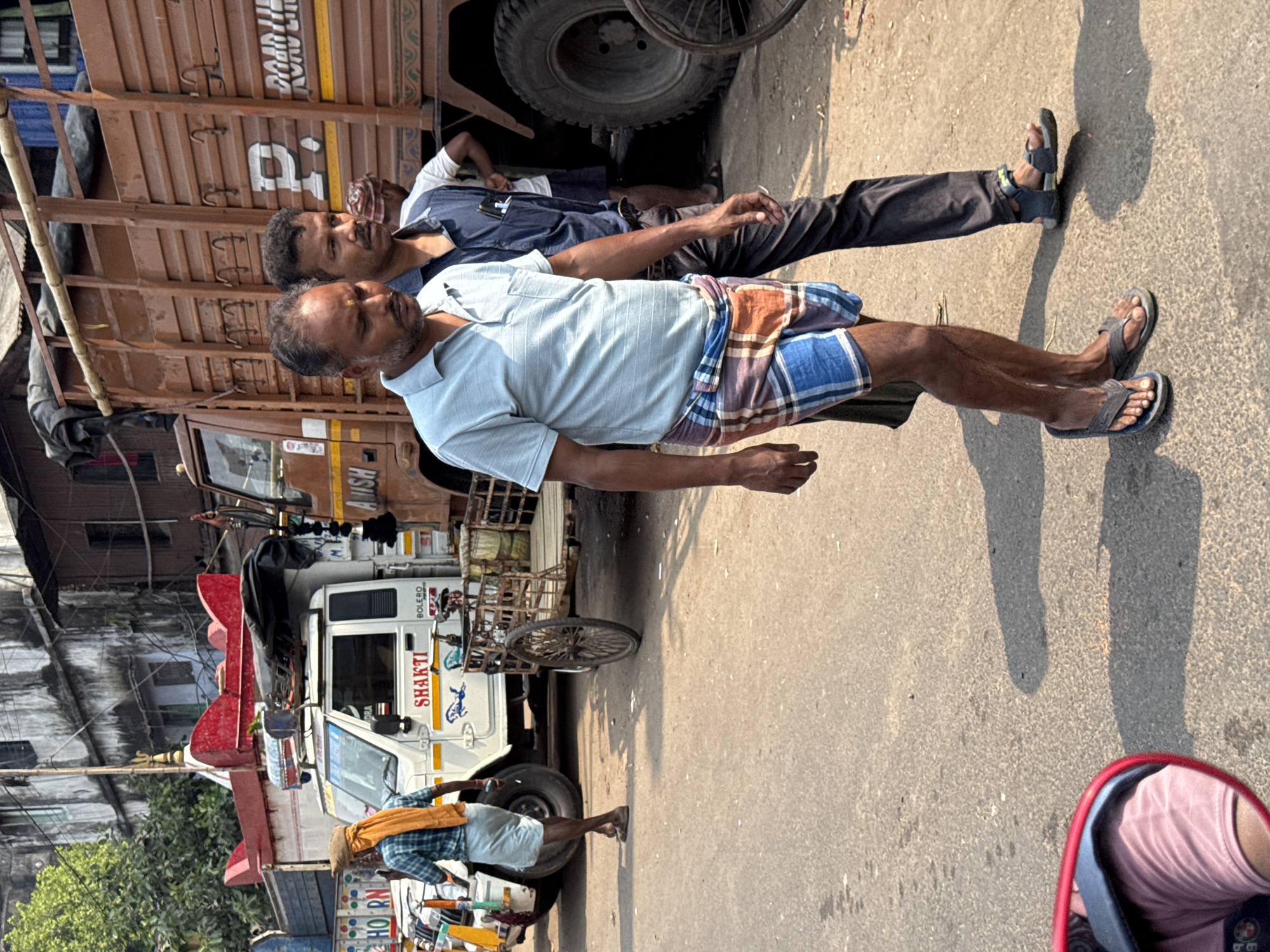 This image depicts a bustling street scene in Kolkata, India. The foreground features two adult men walking right to left on an asphalt road. The man on the left wears a light blue striped polo shirt, a multicolored plaid lungi (blue, white, orange, red), and dark flip-flops; a small yellow mark is visible on his forehead. The man to his right, slightly behind, wears a dark blue collared shirt with a pen in its breast pocket, dark trousers, and dark sandals; he looks towards the right side of the frame.
In the midground, a white Mahindra Bolero camper vehicle is visible, facing left. It has "SHAKTI" and "BOLERO" branding, and an ornate, red, multi-tiered structure, possibly religious or ceremonial, is mounted on its roof, topped with a trishul-like emblem. A black tarp covers additional items on the roof rack. To the left of this vehicle, a man in a plaid shirt, light-colored dhoti, and an orange cloth draped over his head and shoulder walks away from the viewer, holding a mobile phone.
Further to the right in the midground, a large brown heavy-duty truck with wooden slat sides is partially visible, displaying "ROAD LINE" and "AUSH" on its body. A man in a white t-shirt and dark shorts, with a red and white patterned cloth on his head, is seen leaning against the truck's large tire. Between the white camper and the brown truck, a two-wheeled, wooden-framed hand-pulled cart is parked.
The background shows dilapidated multi-story urban buildings with visible windows and some green foliage. The asphalt road is worn with minor stains. A red-framed rearview mirror, reflecting a person in a pink shirt, is visible in the lower left corner, indicating the photographer's position from a vehicle. The scene is illuminated by strong sunlight, casting distinct shadows.