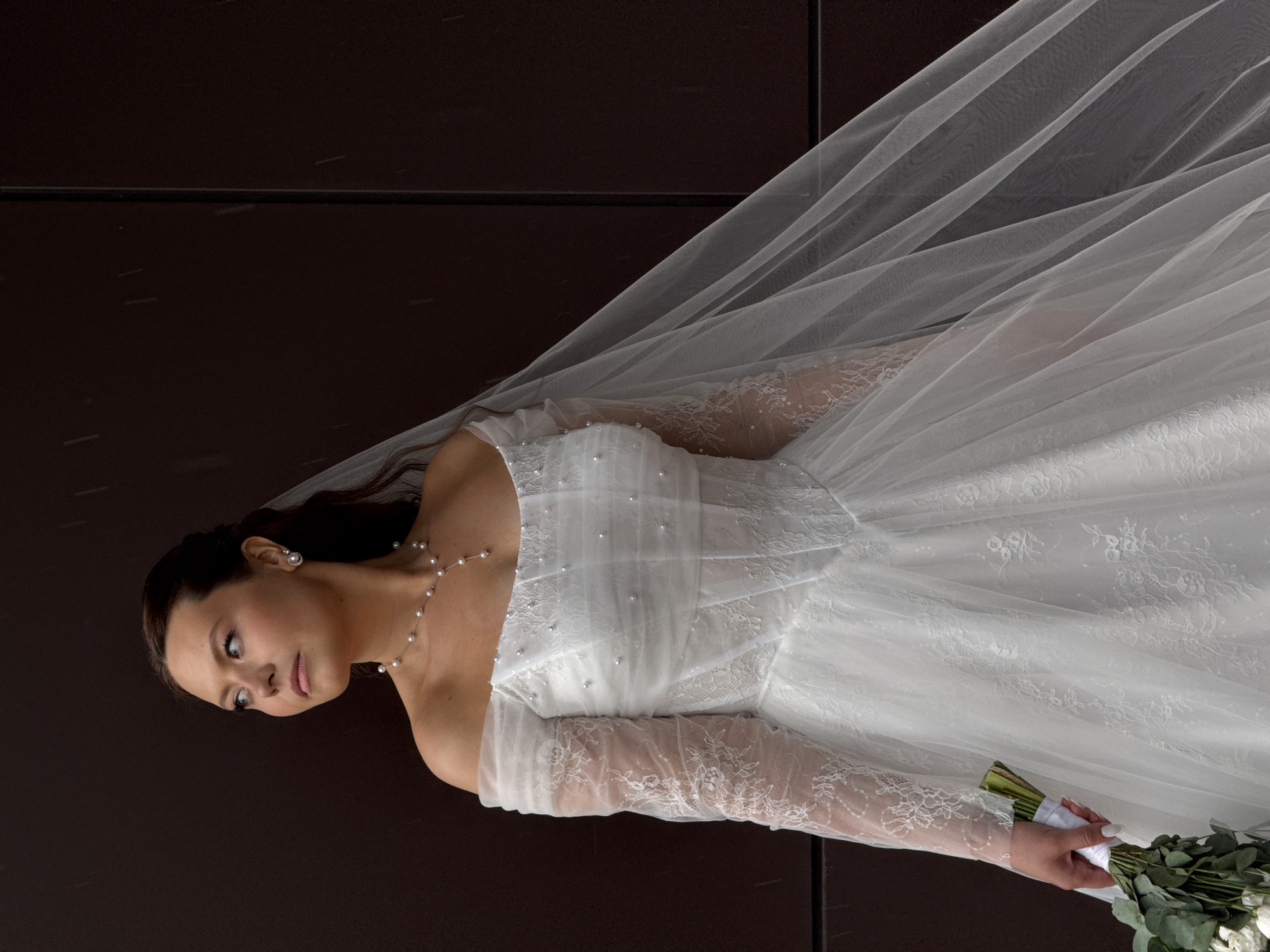 A portrait-style image features a woman dressed as a bride, positioned against a dark brown background. The woman, with dark hair parted to the side, looks gently towards her left, exhibiting a calm and perhaps pensive expression. She is wearing a white, off-the-shoulder wedding gown. The bodice of the dress is embellished with small pearls or beads, and it has long, sheer lace sleeves that extend to her wrists. A delicate pearl necklace adorns her neck, and she wears matching pearl stud earrings. A long, flowing white veil billows behind and to her right, creating soft, translucent layers. In her left hand, she holds a bouquet composed mainly of green foliage, possibly eucalyptus, intertwined with some white flowers. Her fingernails are painted white. The background is a solid, dark brown color, possibly a wall or panel, featuring a subtle horizontal line and minimal light speckles that could be dust or reflections. There is no visible text in the image.