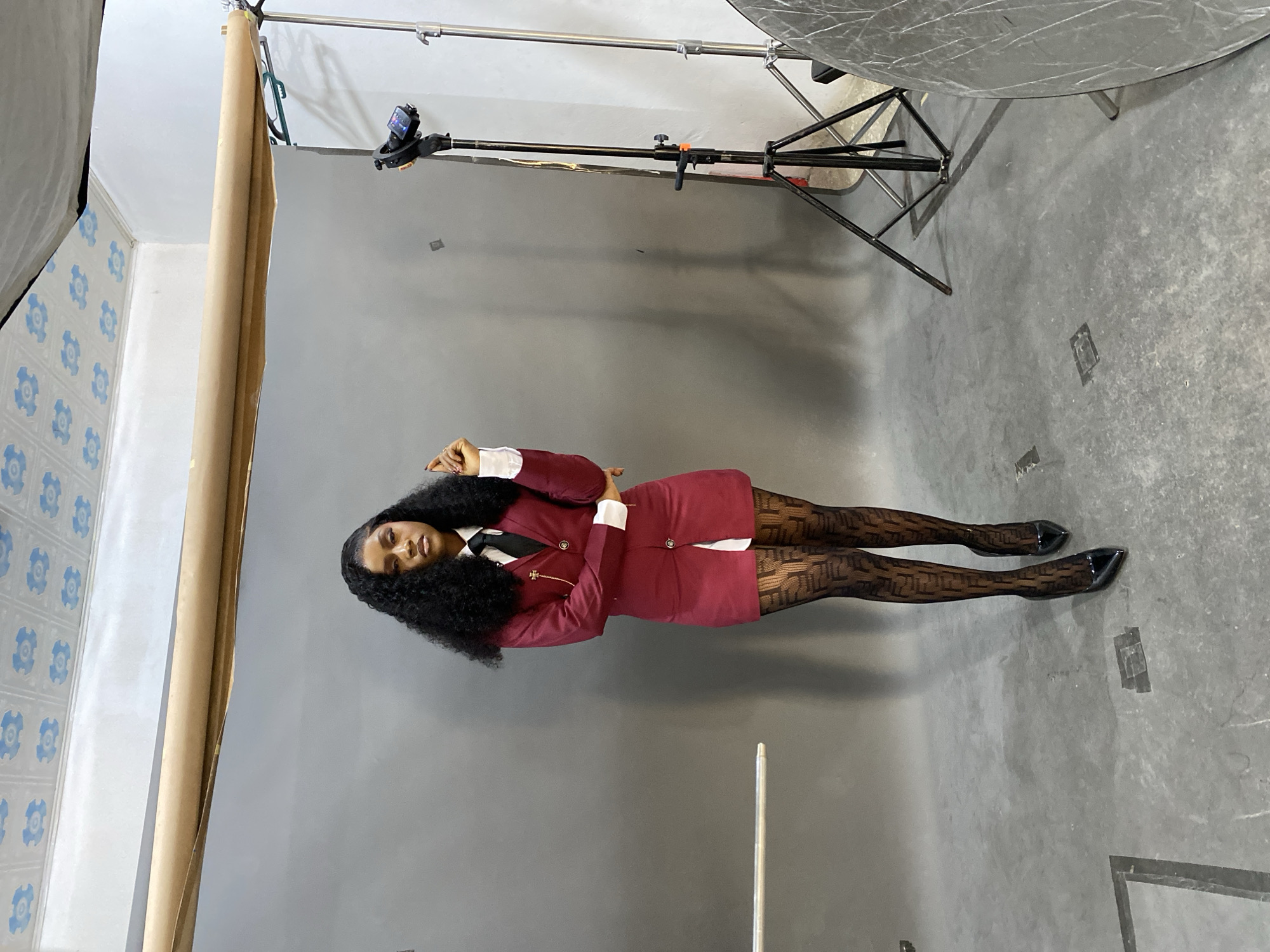 The image displays a young Black female model posing in an indoor photography studio located in Gura Top, Nigeria. She stands centrally, facing the camera with a direct, serious gaze. Her dark, voluminous curly hair falls to her shoulders. She wears a dark red blazer dress featuring white cuffs, a white collar, and a black tie. A gold-toned lapel pin with a cross design and a small chain is affixed to her left lapel. She is clad in black patterned sheer stockings and black pointed-toe high heels. Her left hand is raised slightly, fingers curled, near her chest, while her right arm is bent with her hand resting near her waist. The backdrop is a seamless roll of gray paper, draped over a metal support system. The floor is gray concrete, exhibiting minor scuff marks and two square metal utility covers. The studio walls are white, with a horizontal silver bar mounted on the left wall. The white ceiling features a repeating pattern of blue floral or geometric motifs. To the right of the model, various studio equipment is visible, including a tall black light stand with a camera or monitor mounted at its top, a silver light stand, and a large circular silver reflector resting on the floor.