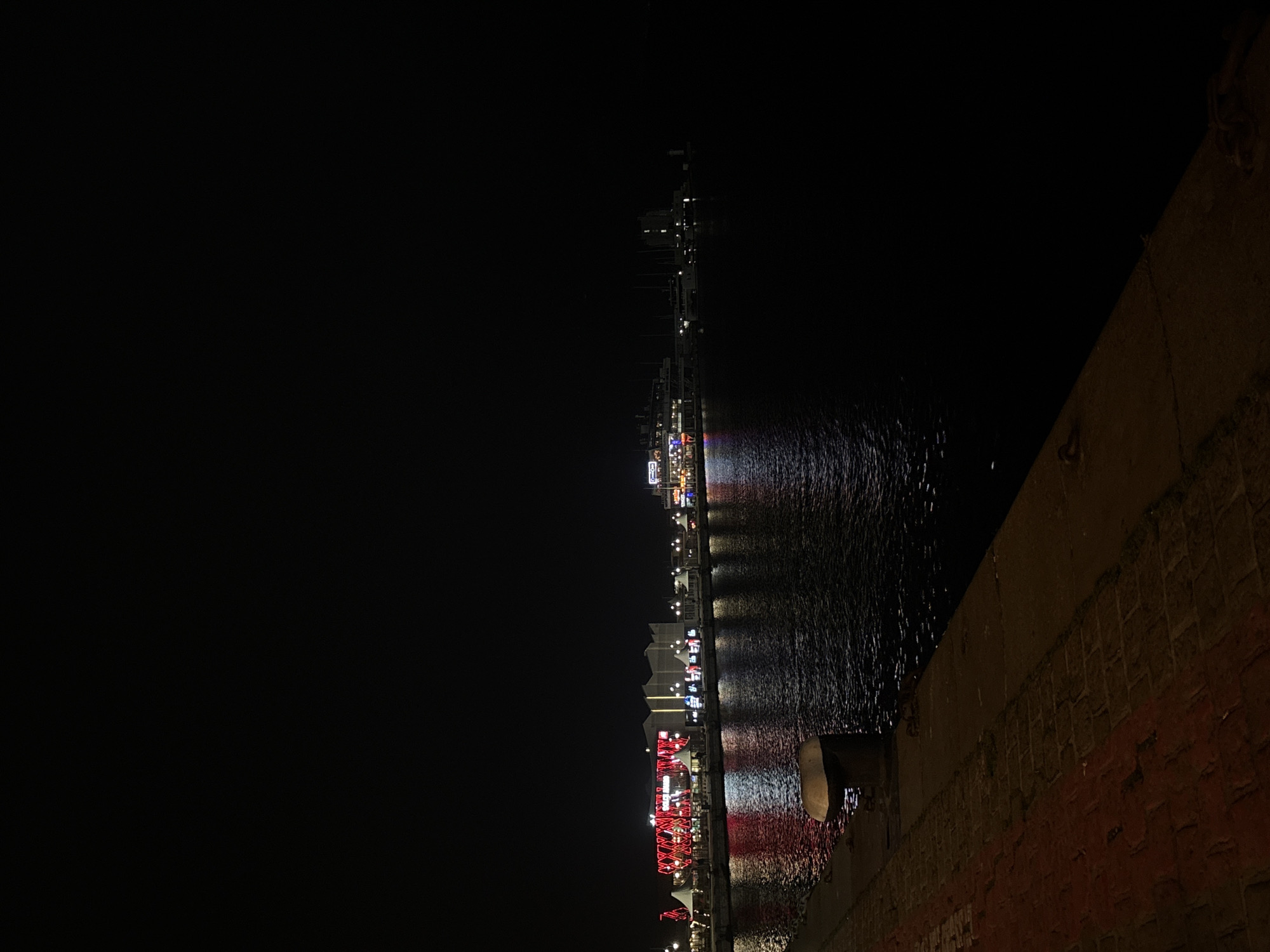The image depicts a night scene in Varna, Bulgaria, showing a waterfront area. A concrete or brick wall runs along the right side of the image. To the left of the wall is a body of water, presumably the Black Sea. Reflected in the water are bright lights from a pier or boardwalk structure extending into the water. The pier is illuminated with red and white lights. The left side of the image is dominated by darkness, suggesting the night sky. A communication mast is barely visible on the pier. The city lights reflect off the water surface.