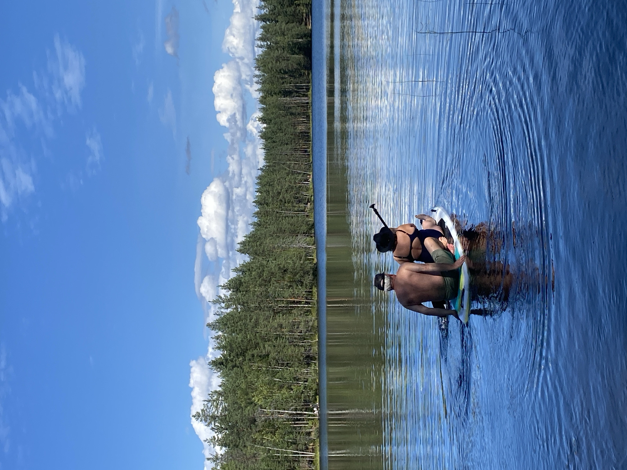 In Leipyasuo, Russia, two individuals are seated on a paddleboard in a tranquil lake. The man, facing away from the camera, is shirtless and wears green shorts and a white baseball cap. The woman, also facing away, is in a dark swimsuit and a wide-brimmed hat. They are positioned on the paddleboard, with the woman holding a paddle. The lake's surface is calm with ripples emanating from the paddleboard. A dense forest of pine trees lines the opposite shore, with a bright blue sky and scattered white clouds overhead. The scene is bathed in natural sunlight.