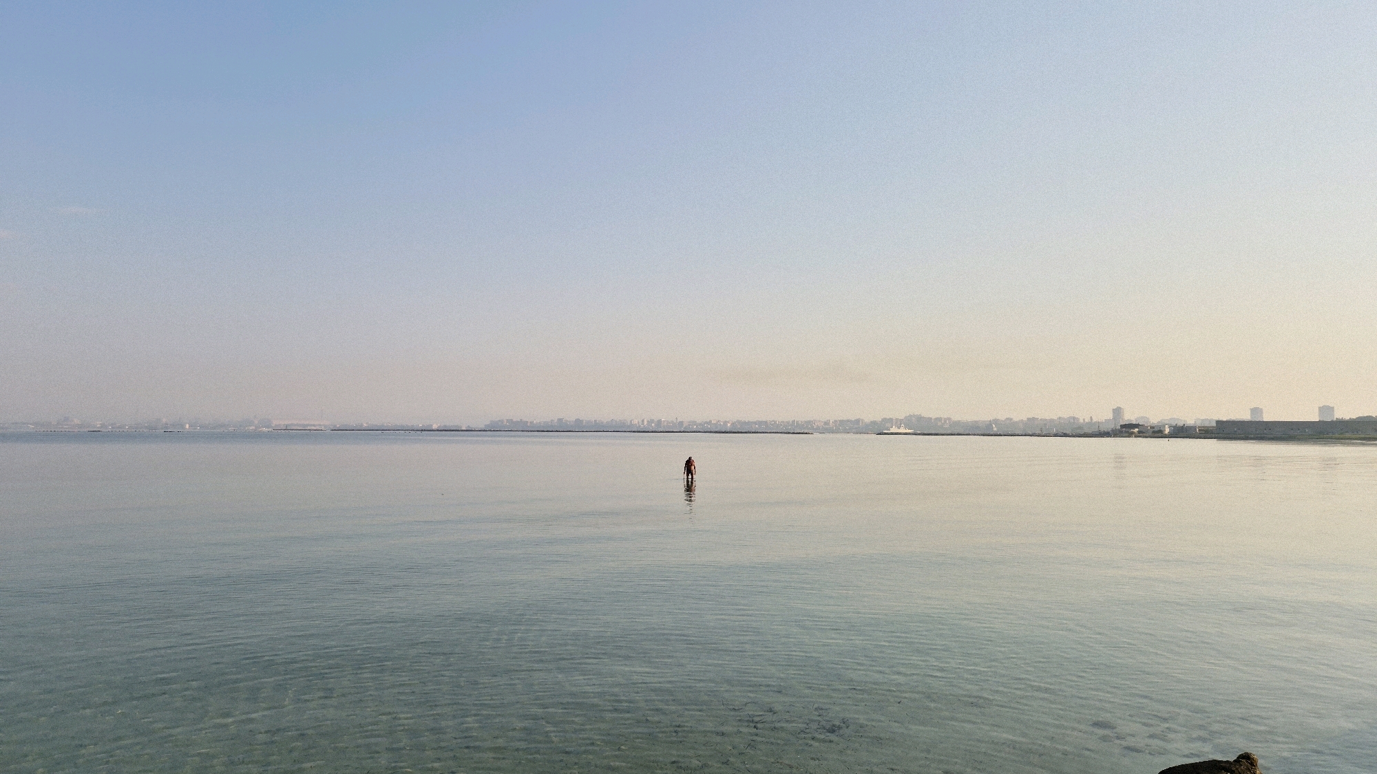 A lone figure stands in the shallow water of the Mar Piccolo, a body of water bordering the city of Taranto, Italy. The figure, appearing to be a male, is wearing dark shorts and a red shirt, and is facing away from the camera. The water is calm and reflects the clear, pale blue sky. In the distance, the skyline of Taranto is visible, a hazy silhouette of buildings and industrial structures. The horizon is a thin line separating the water from the city. The water in the foreground is transparent, allowing the sandy bottom to be seen. The overall atmosphere is one of stillness and solitude.