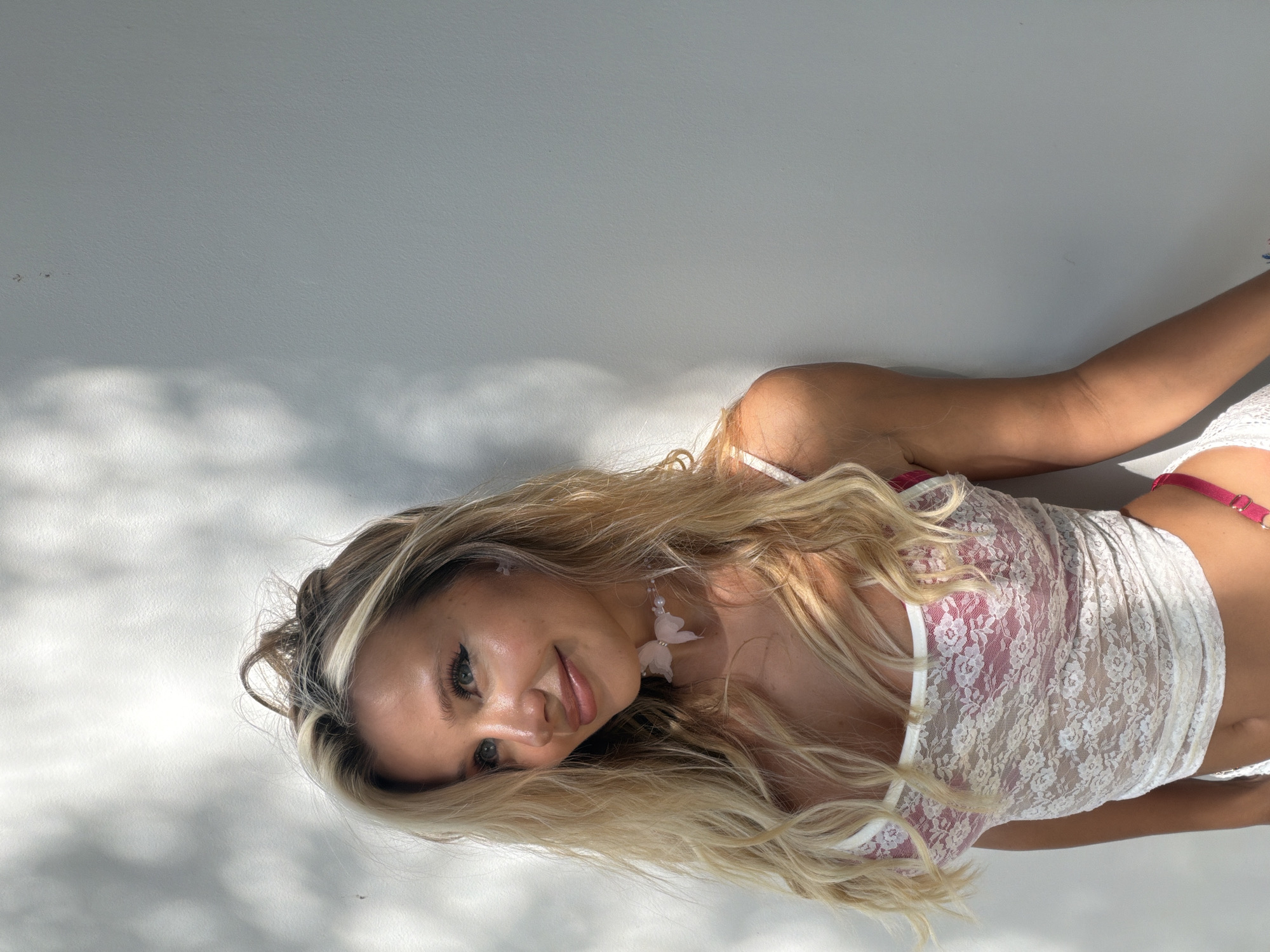 A young woman with blonde hair and blue eyes is posing against a plain white wall. She is wearing a white lace top with a pink bra underneath and pink bottoms. She has a delicate pink choker around her neck. The scene is lit by sunlight, which casts shadows on the wall behind her. The woman is looking off to the side with a slight smile. The overall impression is one of a casual, outdoor portrait. The background does not contain any specific landmarks or features that would indicate a location within Thep Krasatti, Thailand.