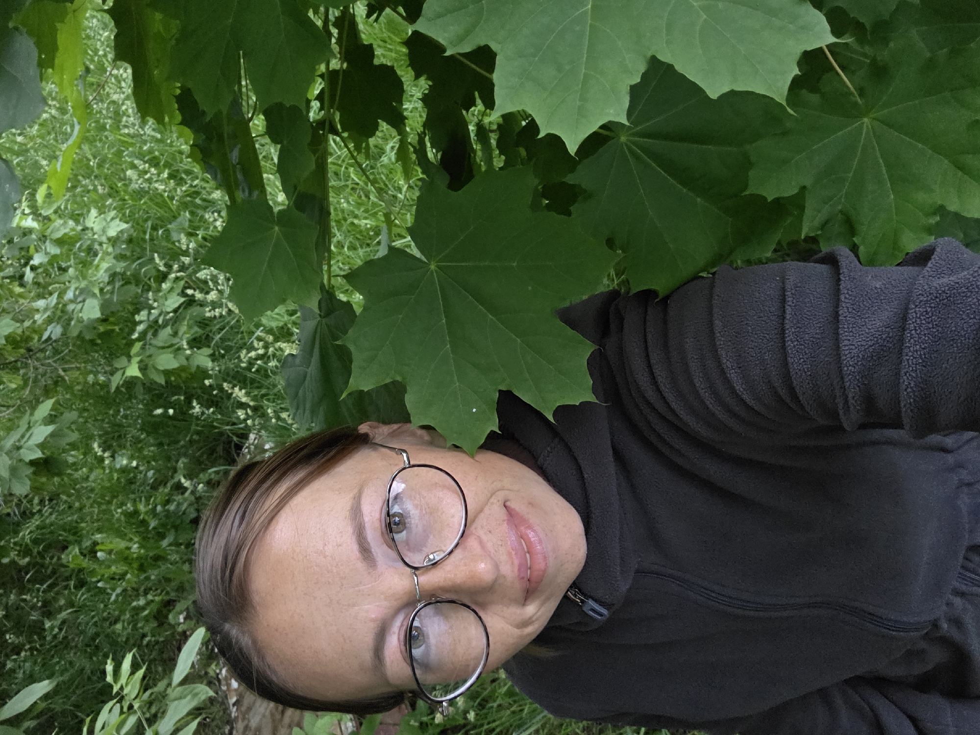 A woman is depicted in a close-up, upward-angled shot in a verdant outdoor environment within the city of Chaikovsky, Russia. She possesses medium-brown hair, pulled back, and her face exhibits freckles. She wears round, metallic-rimmed eyeglasses and displays a faint smile with slightly parted lips. Her gaze is directed straight at the camera. She is clad in a dark-colored, textured fleece jacket or hoodie with a central zipper. The background is characterized by dense green foliage, featuring prominent large, palmate leaves (consistent with maple leaves) in the right foreground and mid-ground. Behind these, a mix of lighter green grass and small leafy plants with scattered white flower clusters is visible, indicating a natural, overgrown setting.