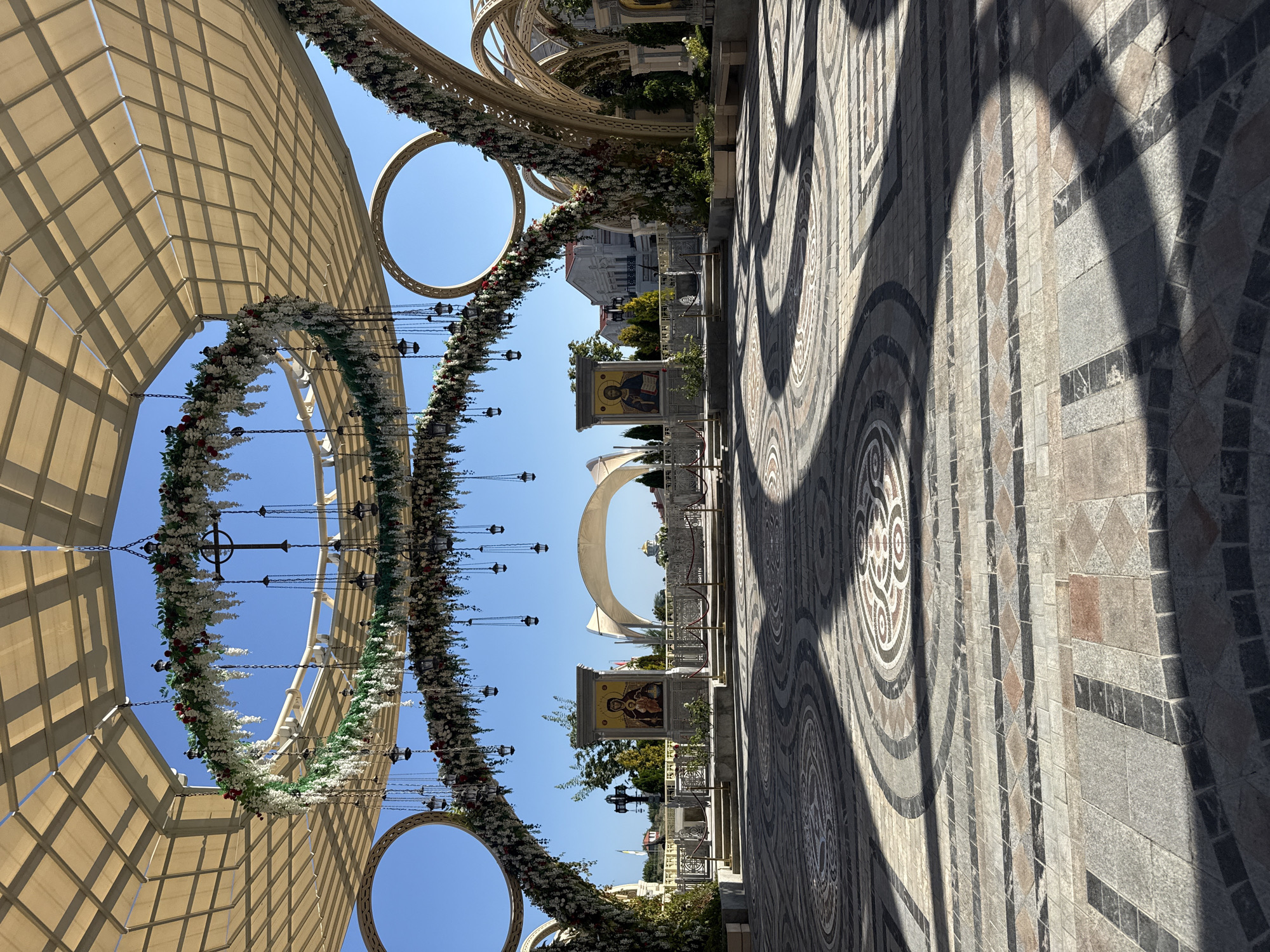 The image shows a wide, paved courtyard decorated with ornate floral garlands and hanging lanterns under a large, open-air canopy. The courtyard is tiled with intricate circular patterns and rectangular stone blocks, with strong shadows cast across it. In the background, there are several buildings, including what appears to be a church with a golden dome, and a large archway. Two icons, one depicting the Virgin Mary and child and the other depicting Jesus Christ, are prominently displayed on pedestals. The scene suggests a religious or ceremonial site. This is the entrance to the Church of Saints Peter and Paul in Sevastopol, Ukraine.