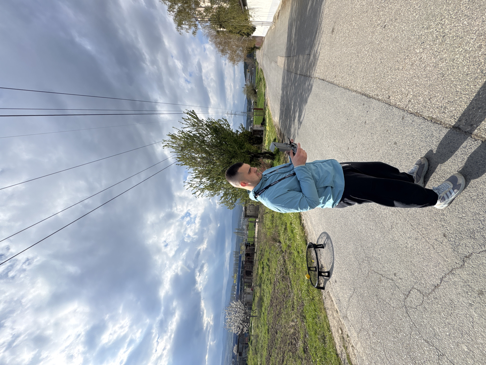 A man stands on a paved road, holding a remote control. He wears a light blue jacket, dark pants, and grey sneakers. Adjacent to the road is a grassy area. In the background, power lines stretch across a cloudy sky. Further in the distance, structures and hills can be seen. Next to him is a ball and a racket. The location is in Blagoevgrad, Bulgaria.