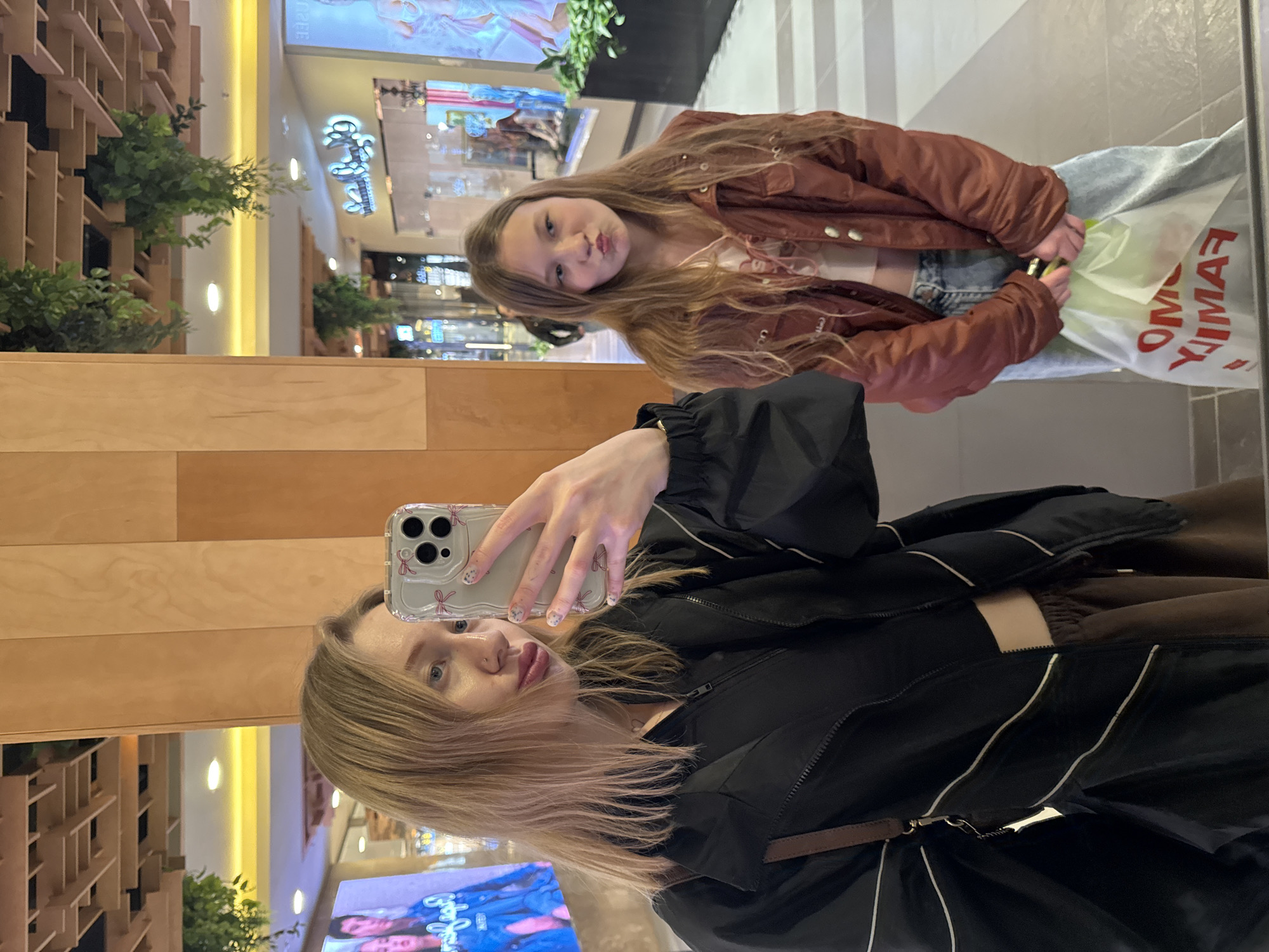 Two females are reflected in a mirror. The taller female holds a smartphone, taking a selfie with the other female in the reflection. The mirror reflects a store within a mall setting. The location of the photo is unknown.