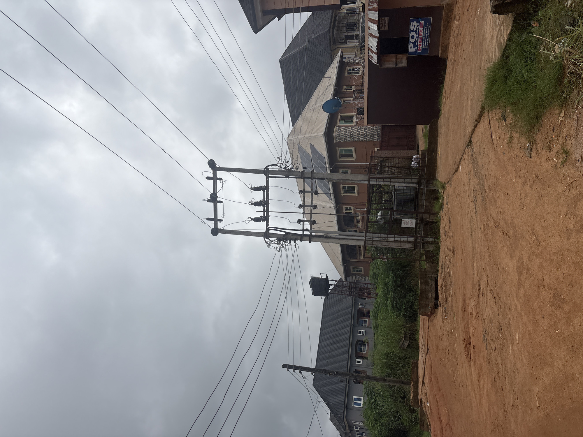 In Obosi, Nigeria, a street scene unfolds under an overcast sky, dominated by electrical infrastructure and residential buildings. A prominent utility pole with multiple crossbars and insulators stands center-frame, supporting a tangle of power lines that crisscross the sky. A large transformer is visible at the base of the pole, enclosed by a metal fence.
To the left, a gray, two-story building with a dark, sloped roof is partially obscured by overgrown green vegetation. It features a series of windows with white frames and a water tank on a tall, metal support structure. Further down the street, another multi-story building, this one with a reddish-brown facade and a light-colored, sloped roof, is visible. This building has solar panels installed on its roof and a satellite dish mounted on its side.
A dirt path, marked by patches of dry, reddish soil and sparse green grass, leads towards the buildings. A small kiosk or shop with a corrugated metal roof is situated on the right side of the scene. A blue sign on the front of the kiosk reads "POS IS AVAILABLE HERE" and lists services such as "Deposit Money," "Withdraw Money," and "Transfer Money." The overall impression is of a developing urban or suburban area with ongoing infrastructure and residential construction.