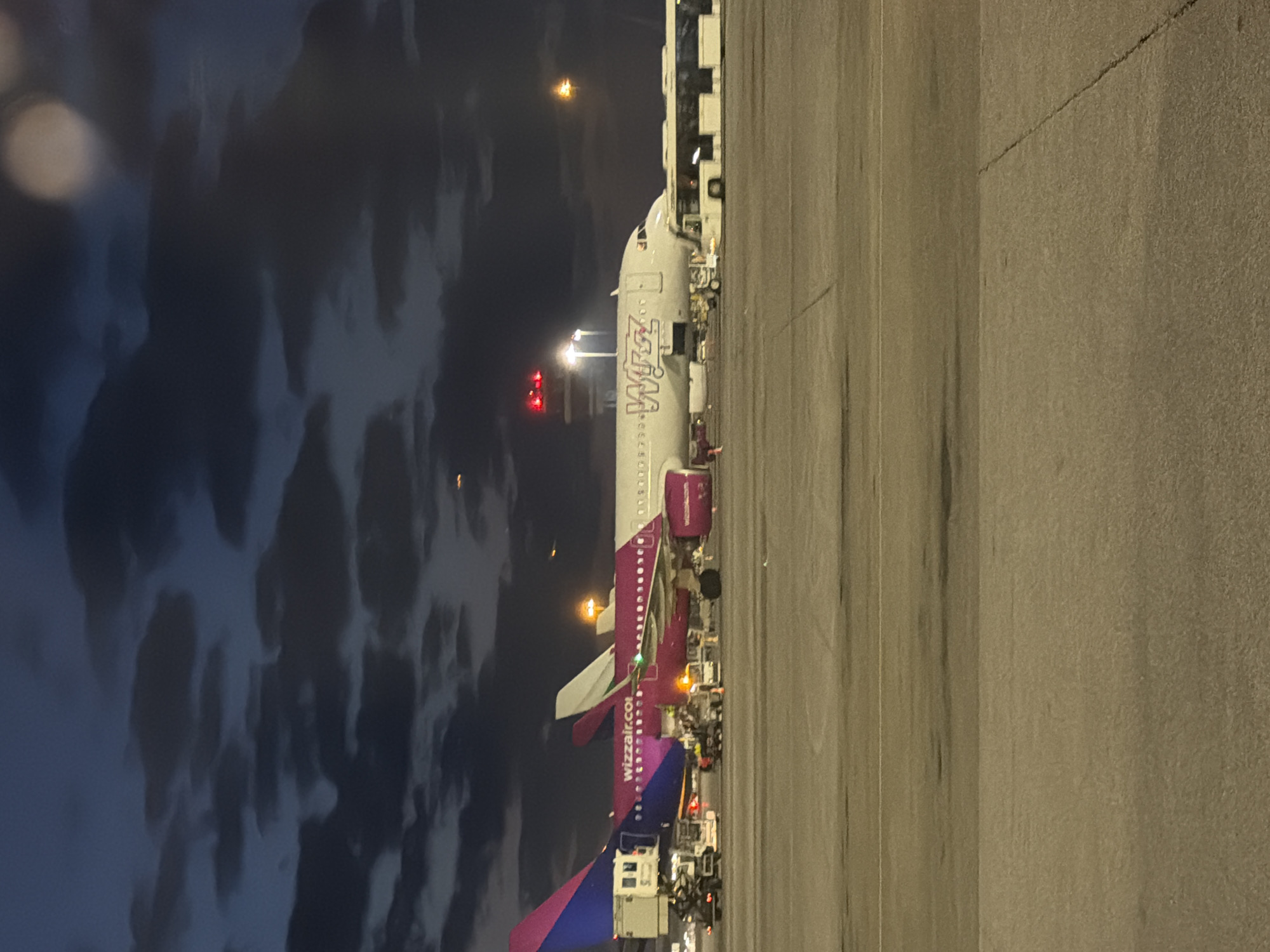 An aerial view looking down at a Wizzair airplane parked on a tarmac at night. The plane is connected to a jet bridge and there are multiple ground vehicles surrounding the plane. The sky is dark with some clouds visible. The location is likely at Sofia Airport in Sofia, Bulgaria, given that Wizzair has a significant presence at that airport.
