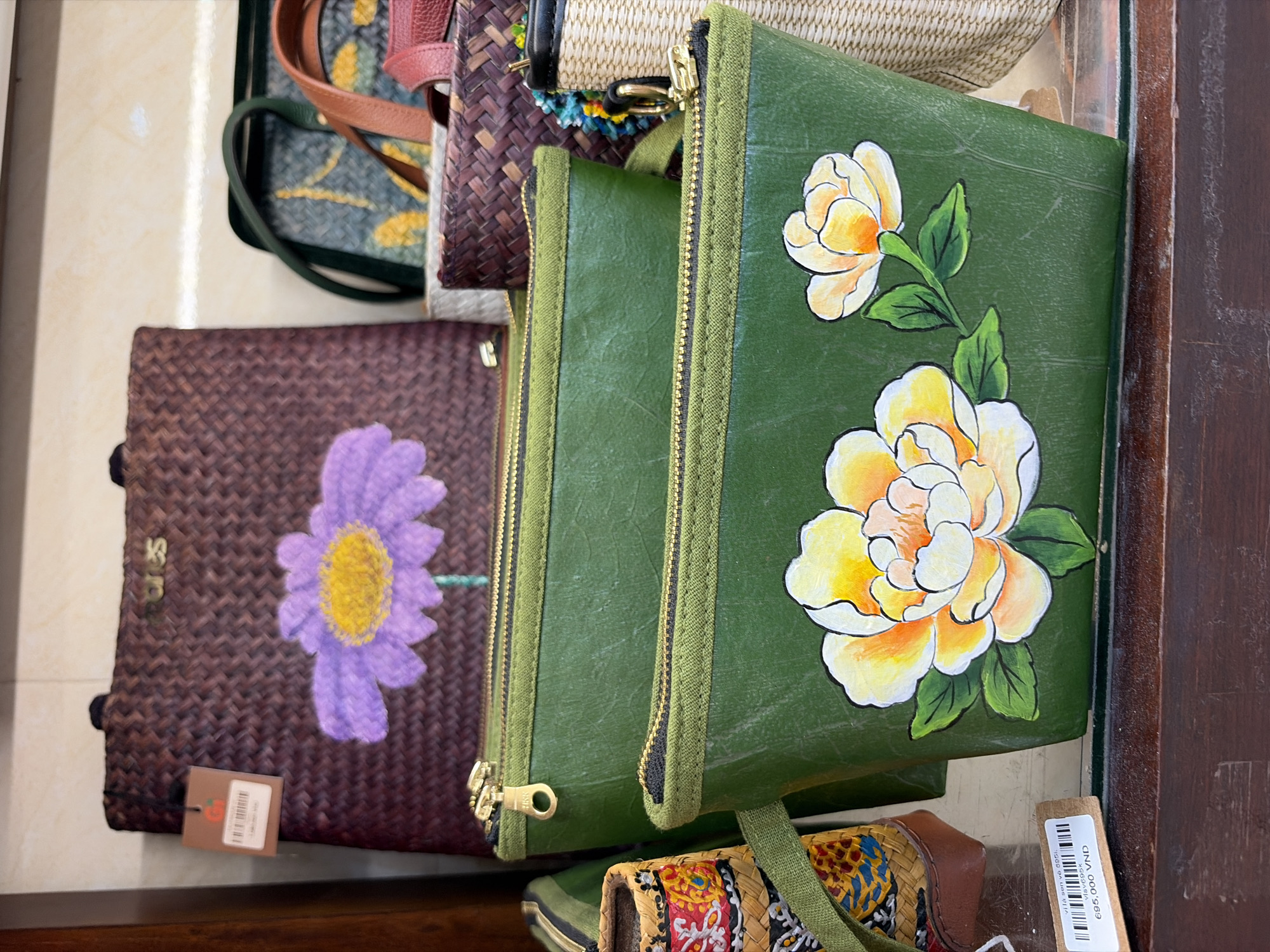 The image shows a close-up of a display of handmade bags, likely in a shop in Da Nang, Vietnam. In the foreground, two green zippered pouches with hand-painted flowers are stacked vertically. The bottom pouch features a large yellow and white peony with green leaves, while the upper pouch is partially obscured but appears to be similar in style. The bags are made of a slightly textured material. Behind these pouches, several other bags are visible. A large woven brown tote bag with a painted purple flower is at the top left. To its right, a woven basket bag with green and brown straps is partially visible, and behind that, another woven bag with a colorful pattern. A small price tag with Vietnamese text and "695,000 VND" is attached to the bottom left pouch. The background consists of a light-colored wall and a dark wooden surface, suggesting a retail setting.