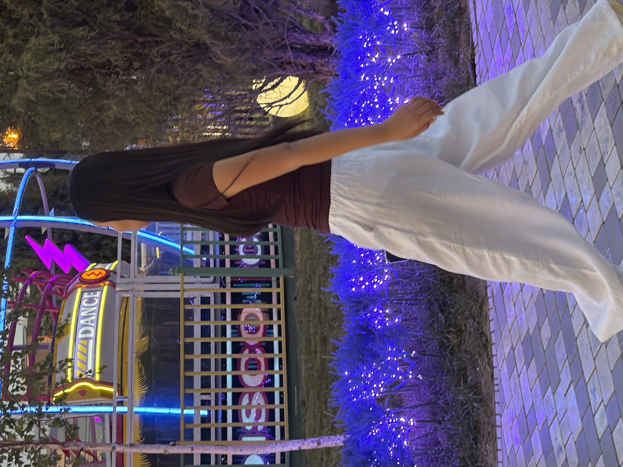 An image taken at night shows a young woman with long dark hair, wearing a maroon t-shirt and white pants, walking from left to right. The background features a vibrant amusement park scene with bright neon lights, including a sign that reads "DANCE" and another that appears to say "DISCO". The ground is paved with gray and purple interlocking bricks. In the foreground, rows of bushes are illuminated with numerous small blue lights, creating a sparkling effect. A thin tree trunk with white bark is visible on the left side of the frame. The overall atmosphere suggests a lively nighttime entertainment venue.