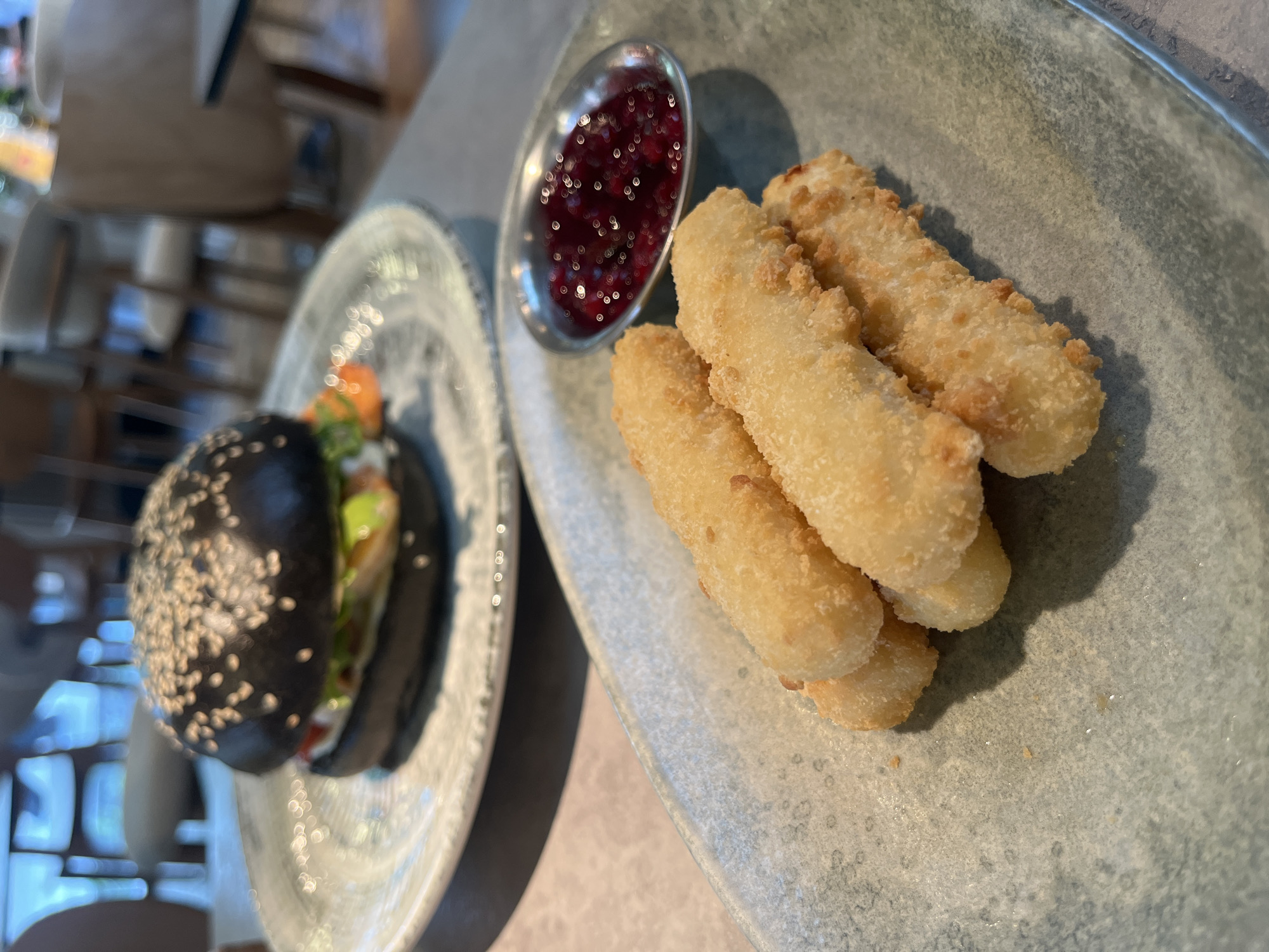 On a textured, light-grey plate, a cluster of golden-fried sticks sits alongside a small, silver dish filled with a dark red jam or sauce. Adjacent, another textured, light-grey plate holds a black sesame seed burger. The scene is captured in daylight, possibly indoors. The location is Unknown, Unknown.