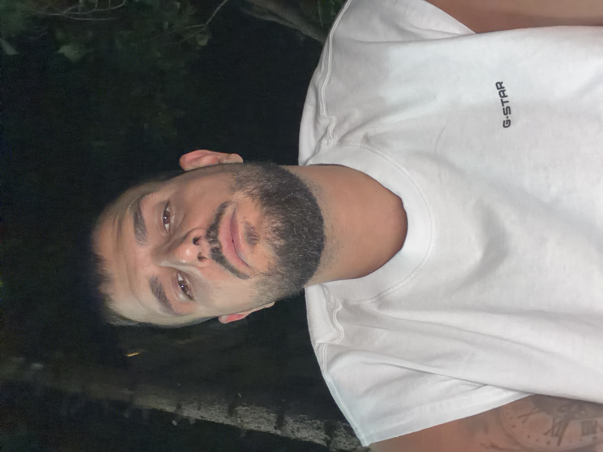 The image shows a close-up, waist-up portrait of a man with short dark hair, a beard, and a mustache. He is wearing a white sleeveless t-shirt with the "G-STAR" logo visible on the left side of the chest. The background is dark and appears to be outdoors at night, with hints of trees and foliage. There is a faint tattoo on the man's left arm, partially visible, that resembles a clock face with Roman numerals. The lighting is dim, casting shadows and highlighting the man's features. There is no discernible location in the city of Sofia, Bulgaria, indicated in the image.