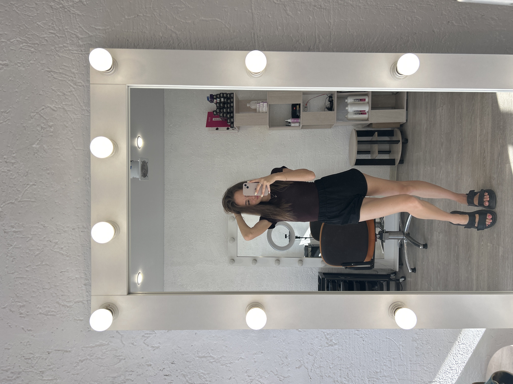 A young woman stands in front of a large, white-framed mirror with lights. She is wearing a dark brown short-sleeved top and black shorts. Her right hand is in her hair, and she is holding a smartphone in her left hand, taking a selfie. She is wearing black sandals. The mirror reflects the room behind her, which appears to be a salon or dressing room. Visible in the reflection are shelves with various bottles and products, a rolling chair, a circular ring light on a stand, and a wall with a textured white finish. The flooring is a light grey wood-grain. The overall lighting is bright, likely from the mirror's bulbs and ambient room light.
