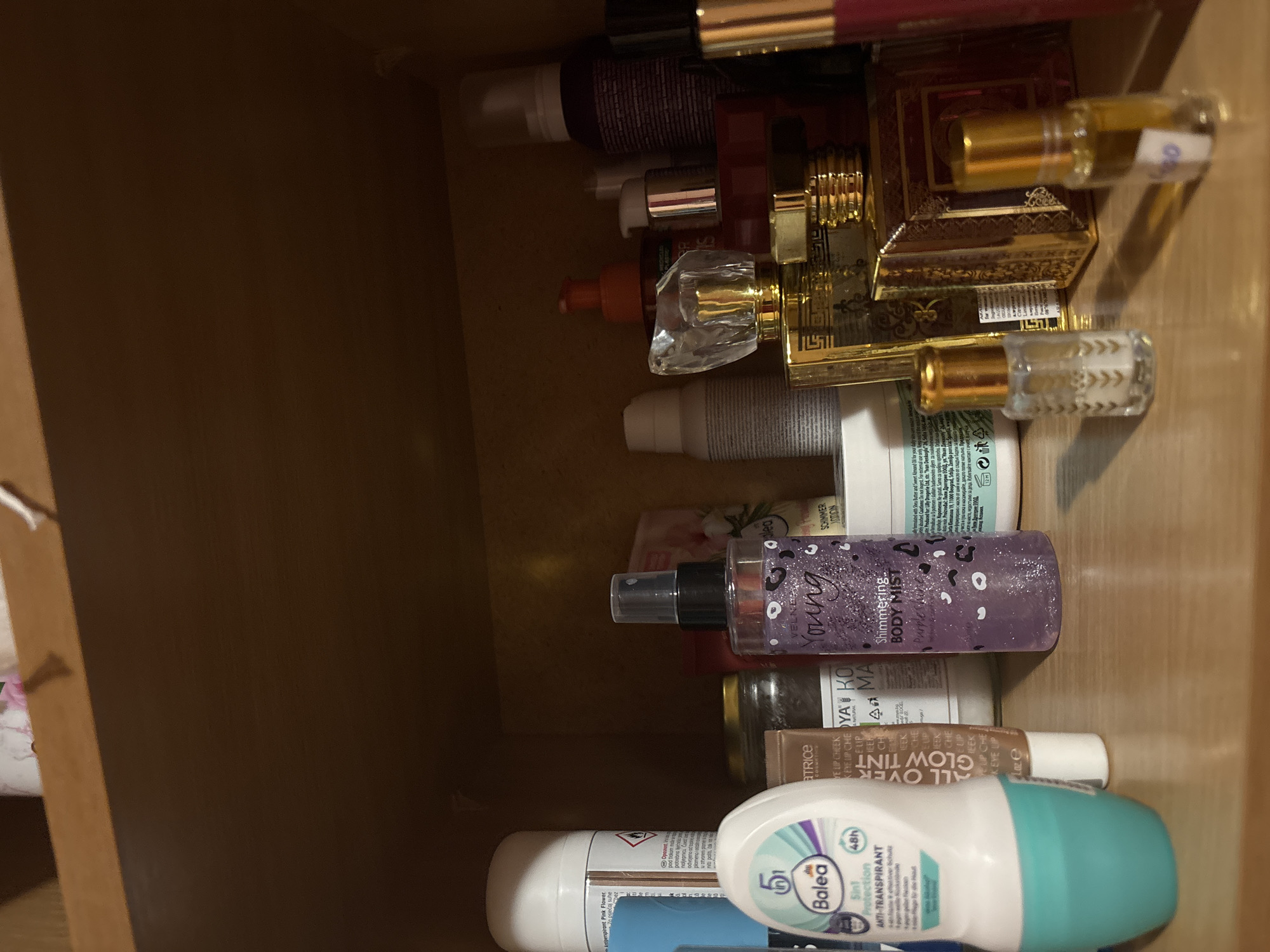 The image shows a collection of beauty and personal care products stored inside a wooden drawer. The products are arranged haphazardly and viewed from an overhead perspective.
Items identified include:
* Cosmetics: Lipsticks or lip glosses (burgundy), gold-tone perfume bottles with intricate designs, an “All Over Glow Tint” product.
* Skincare: A purple “Shimmering Body Mist” by Velnea Young, lotions or creams in various white or beige containers, a product labeled “Koya Ko Ma” and "Kaelp Che."
* Deodorant: A “Balea 5in1 Protection Anti-Transpirant” roll-on deodorant.
The drawer is made of wood, with a medium brown tone, showing the grain. The lighting is dim, with shadows cast within the drawer, making it hard to determine the exact colors and details.
There is no information available to confirm that this photo was taken in Garmen, Bulgaria. The product labeling in the image includes text in Bulgarian, German, and English.