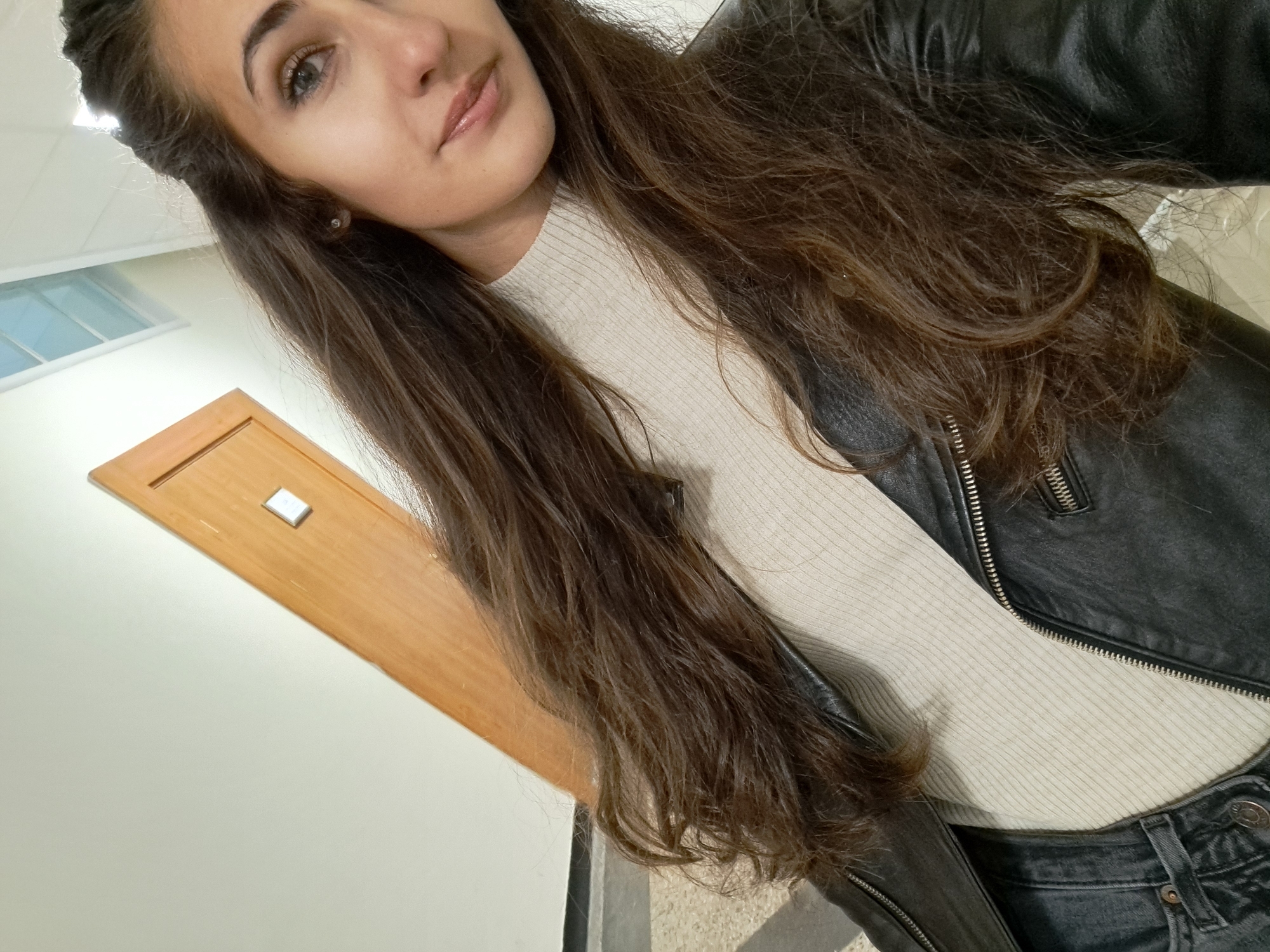 A young woman with long brown hair and wearing a black leather jacket and a light-colored ribbed sweater takes a selfie. The background is an interior setting with light-colored walls and a wooden door. A window is visible in the upper left corner. The photo appears to be taken at a slight angle. The location is unspecified and unknown.
