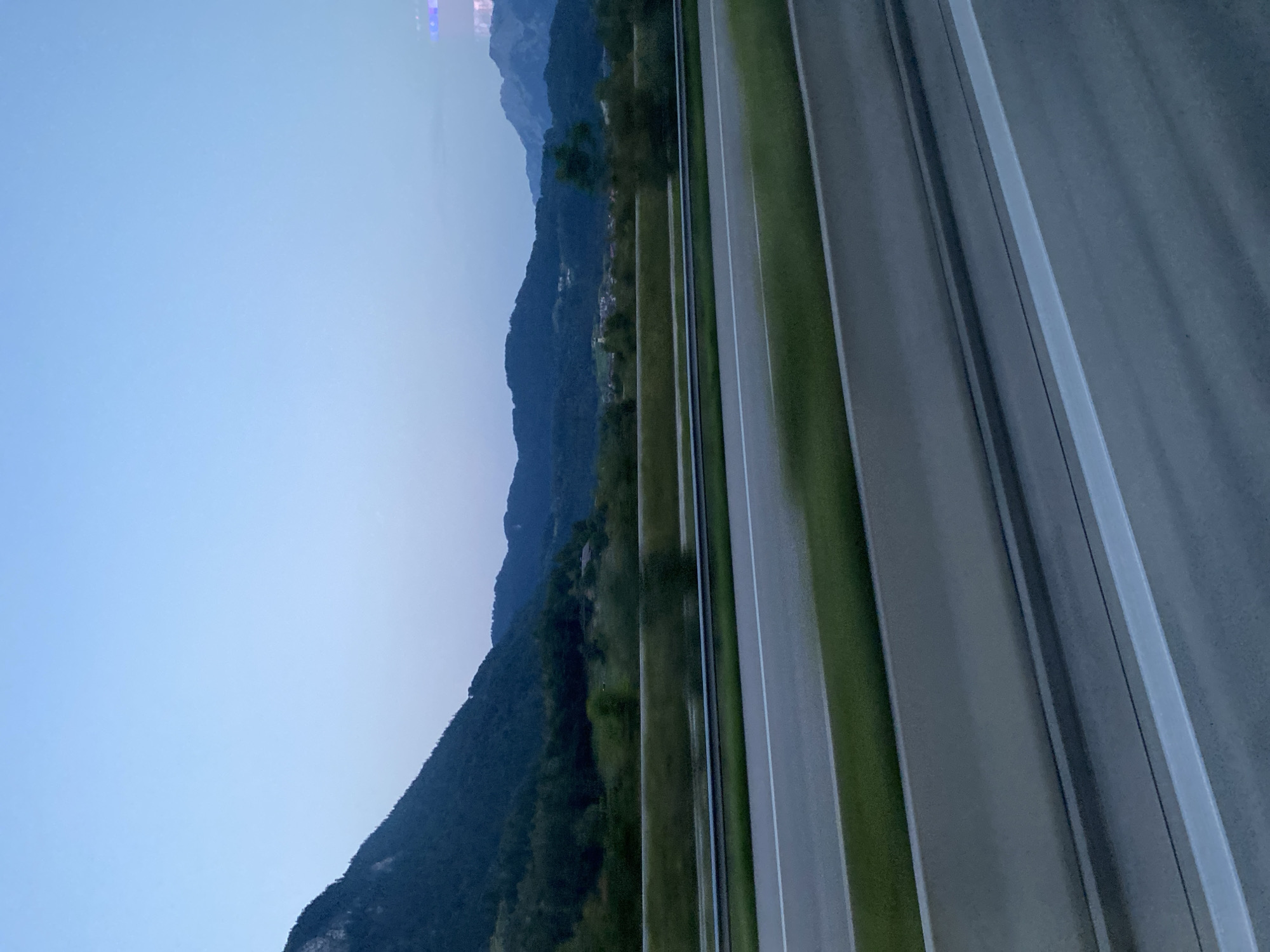 This image depicts a serene landscape near the city of Oberaudorf, Germany, captured during the twilight hours. The foreground features a highway with multiple lanes, distinguished by their grey asphalt and white lane markings. A concrete barrier separates the lanes, and beyond that, a grassy verge with a metal guardrail is visible. The middle ground showcases a lush green landscape with trees and vegetation, gently rolling hills, and in the distance, the majestic, tree-covered mountains that characterize the region. A small village with buildings is nestled within the valley between the hills and mountains. The sky above is a gradient of pale blue, transitioning to a deeper shade as it meets the mountain horizon. The overall atmosphere is peaceful and tranquil, indicative of a rural setting with a well-maintained road infrastructure.