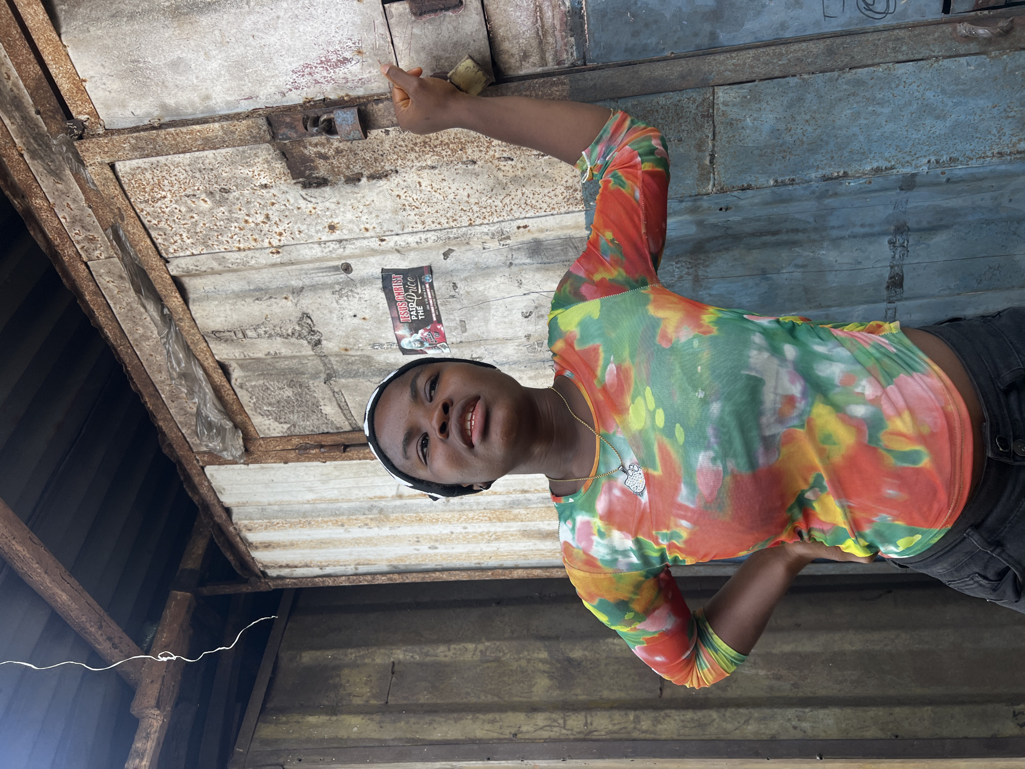 A medium-shot, rotated image depicts a person with brown skin wearing a colorful long-sleeved shirt and dark pants, standing within a dilapidated, corrugated metal structure. The person has a white headband. The structure's roof and walls exhibit rust, dirt, and patches of corrosion. A tattered advertisement is affixed to the ceiling. The location is identified as Unknown, Unknown.