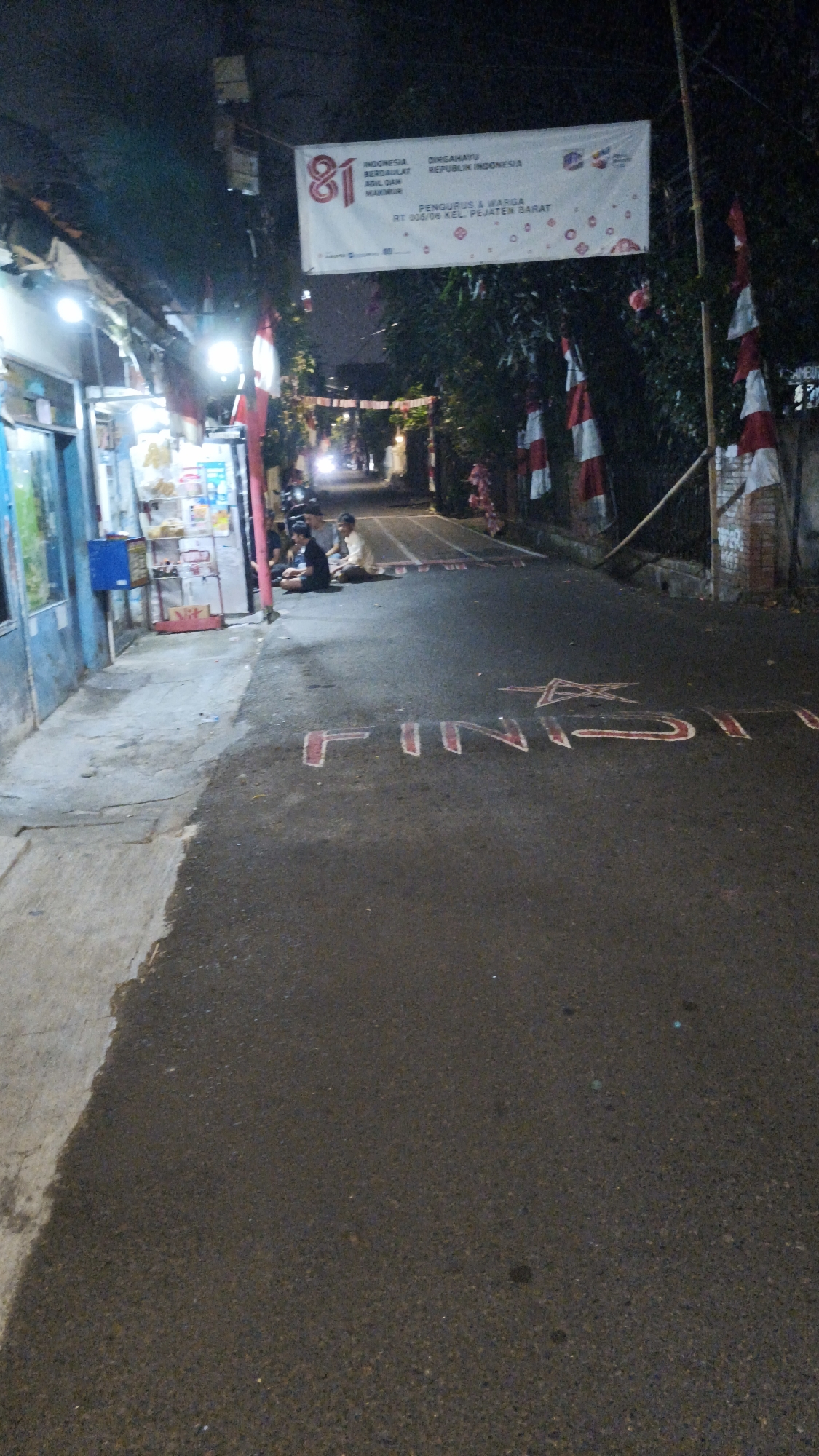The image depicts a nighttime street scene in South Jakarta, Indonesia. A banner overhead reads "DIRGAHAYU REPUBLIK INDONESIA" and "INDONESIA BERDAULAT ADIL DAN MAKMUR" with the number "81" prominently displayed, indicating a celebration of Indonesia's 81st Independence Day. The banner also lists "PENGURUS & WARGA RT 005/06 KEL. PEJATEN BARAT".
The street is paved and appears to be a residential or local commercial area. Several people are seated on the ground near a small shop on the left side of the road, illuminated by the shop's lights. Another group of individuals is further down the street. Red and white Indonesian flags are visible, attached to poles and strung along the street, contributing to the festive atmosphere. The road surface itself has red painted markings, including the word "FINISH" and a star graphic. The overall environment is dimly lit, with artificial lighting from the shop and distant headlights creating shadows and highlights.
