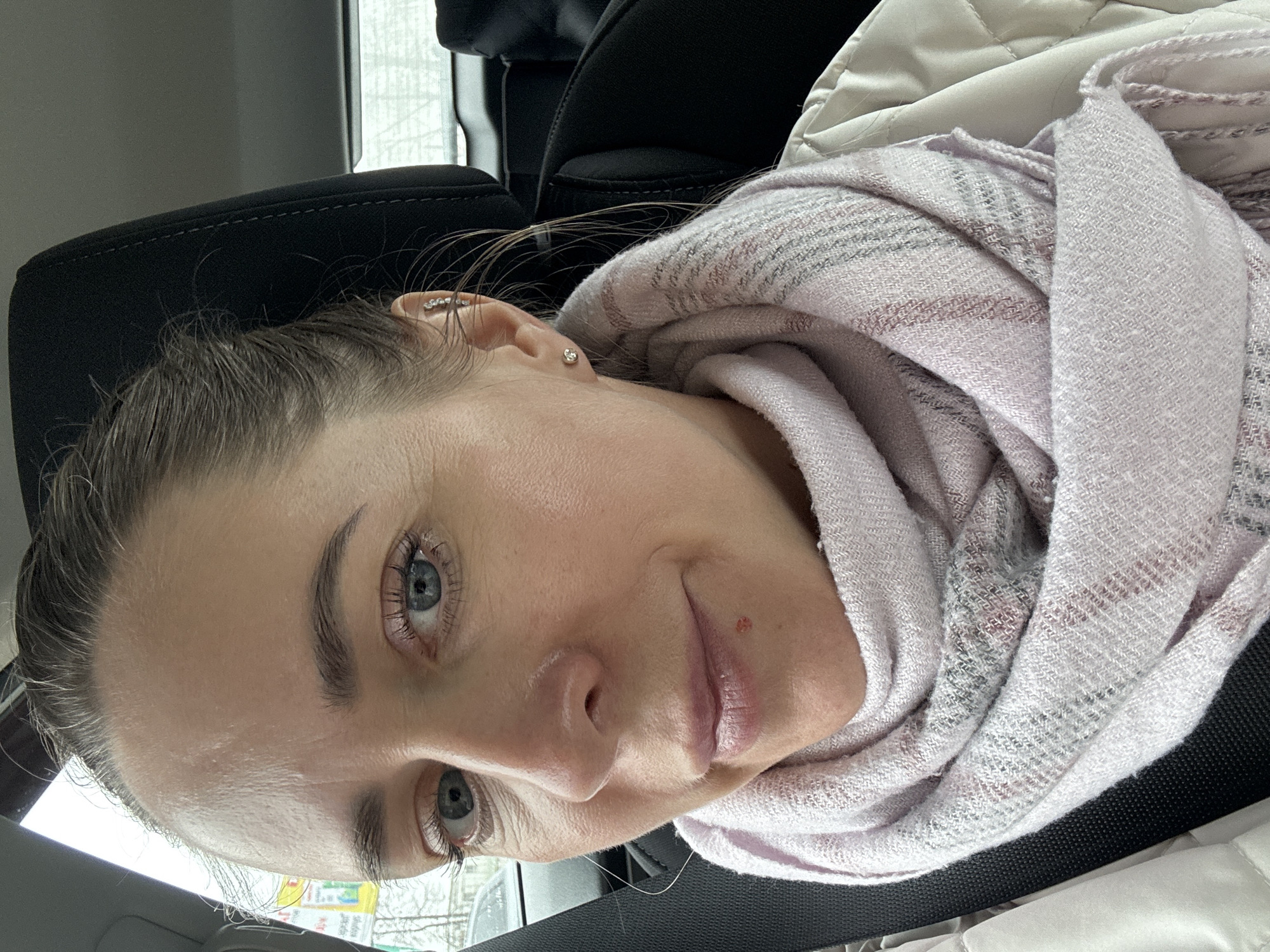 A woman with fair skin and blue eyes is the subject of this close-up, tilted image. She has light brown hair pulled back and is wearing a pale pink and grey scarf. She also has a small red mark near her lip and multiple piercings on her left ear. She is seated in what appears to be a car, indicated by the black car seat behind her and part of a seatbelt across her chest. The environment visible through the car window appears to be an urban setting in Chaikovsky, Russia, with blurred outlines of buildings and potentially wintery conditions outside.