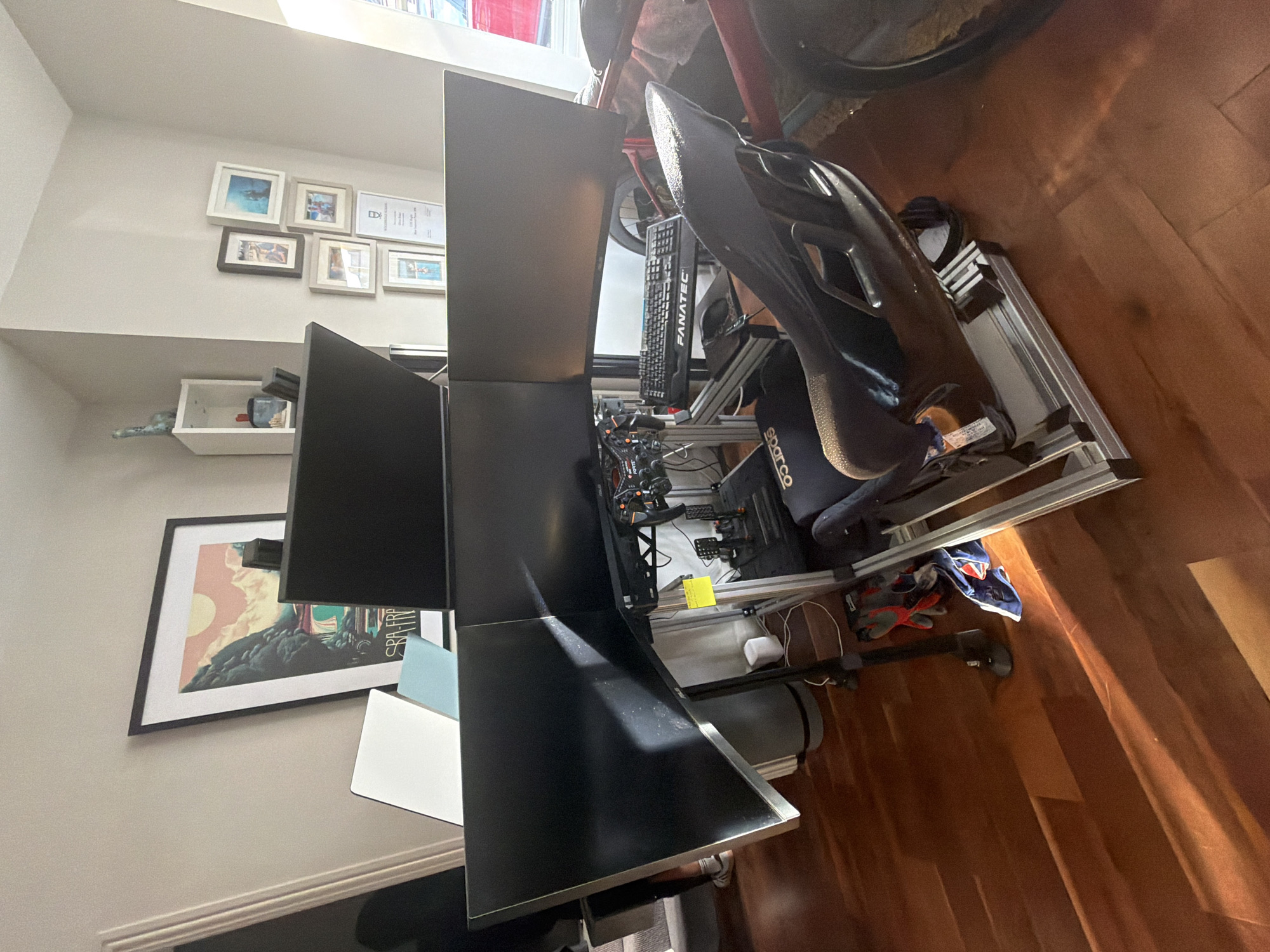 The image shows a gaming setup indoors. A triple monitor display dominates the view, with a "Fanatec" keyboard visible behind it. The setup includes a racing seat with "Sparco" branding, mounted on a metal frame, likely a racing simulator rig. Other components such as a steering wheel and pedals are partially visible.
The room has wooden flooring and light-colored walls. On the wall, there are framed pictures and a small white shelf with items on it. A framed poster is also visible. The location in East Suffolk, UK, cannot be determined from the image.