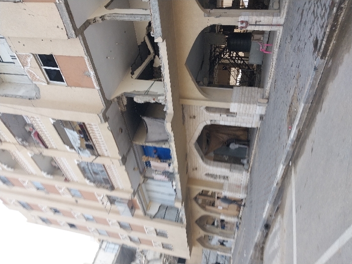 The image captures a severely damaged multi-story building in Khan Yunis, Palestinian Territory, under daylight conditions. The building, constructed with beige-colored exterior walls, shows extensive structural collapse, particularly on its upper floors and façade. Large sections of walls and ceilings are missing, revealing the building's interior framework, including concrete slabs, rebar, and debris. Many windows are broken, shattered, or completely absent.
The ground floor features a series of distinct arched openings. Through one of these arches, a dark, cylindrical tank on a metal structure is visible, along with a pink plastic chair nearby. Another arch reveals a lighter-colored object inside, possibly an appliance or cooler. The area in front of the building consists of paved ground, appearing to be a sidewalk or street made of interlocking pavers and asphalt. No people are visible in the scene, and there is no ongoing activity, only the aftermath of destruction. The overall impression is one of heavy damage, likely from conflict, affecting multiple levels of the urban structure. No visible text is discernible in the image.