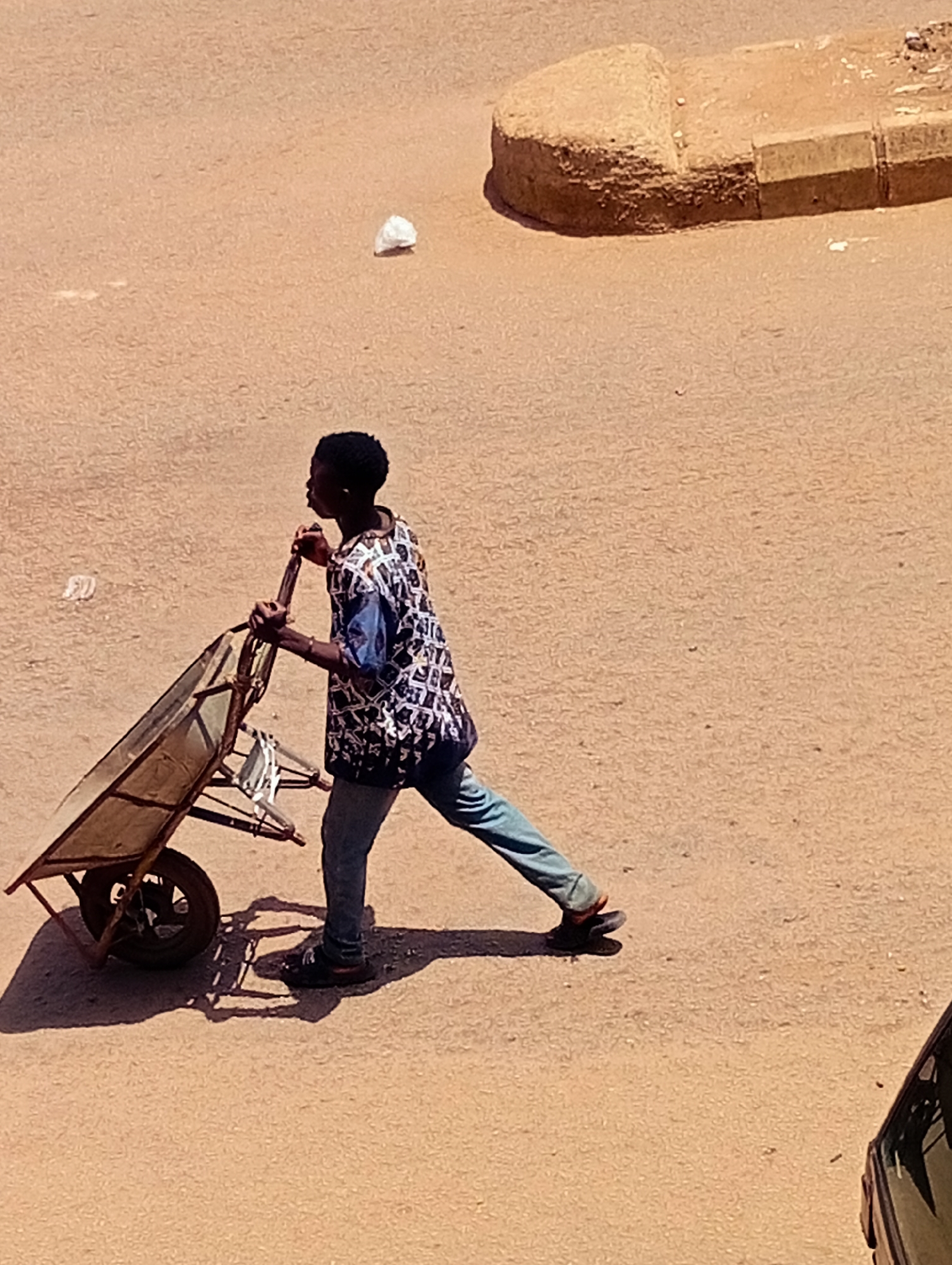 A young Black male, wearing a patterned shirt and blue jeans, pushes a wheelbarrow on a sandy road in Bukuru, Nigeria. The wheelbarrow is made of metal and wood, with a single wheel. The man is walking away from the viewer, towards the right side of the image. A curb made of concrete and dirt is visible in the upper right corner of the image, along with a small white piece of trash. The ground is sandy and appears to be a dirt road or a construction site. A dark car is partially visible in the lower right corner of the image. The lighting suggests it is daytime, with strong shadows cast by the wheelbarrow and the man.