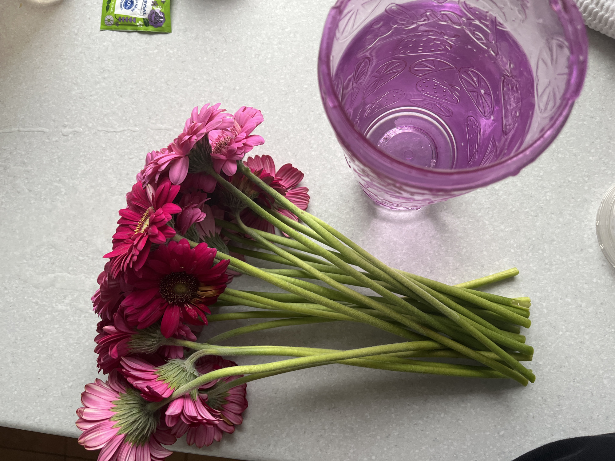 A top-down, medium shot shows a bouquet of pink and magenta gerbera daisies arranged on a light gray, speckled countertop. The flowers are predominantly pink on the left side of the frame and transition to deeper magenta tones on the right. Their green stems are visible, clustered together and slightly splayed. To the right of the flowers, a translucent purple tumbler with a patterned design featuring fruits like strawberries and citrus slices is partially visible, also resting on the countertop. In the upper right corner, a small, green, foil packet, possibly a floral preservative or supplement, is present. The lighting suggests an indoor setting, possibly a kitchen or a general living area. There is no indication of the location within Kohtla-Järve linn, Estonia.