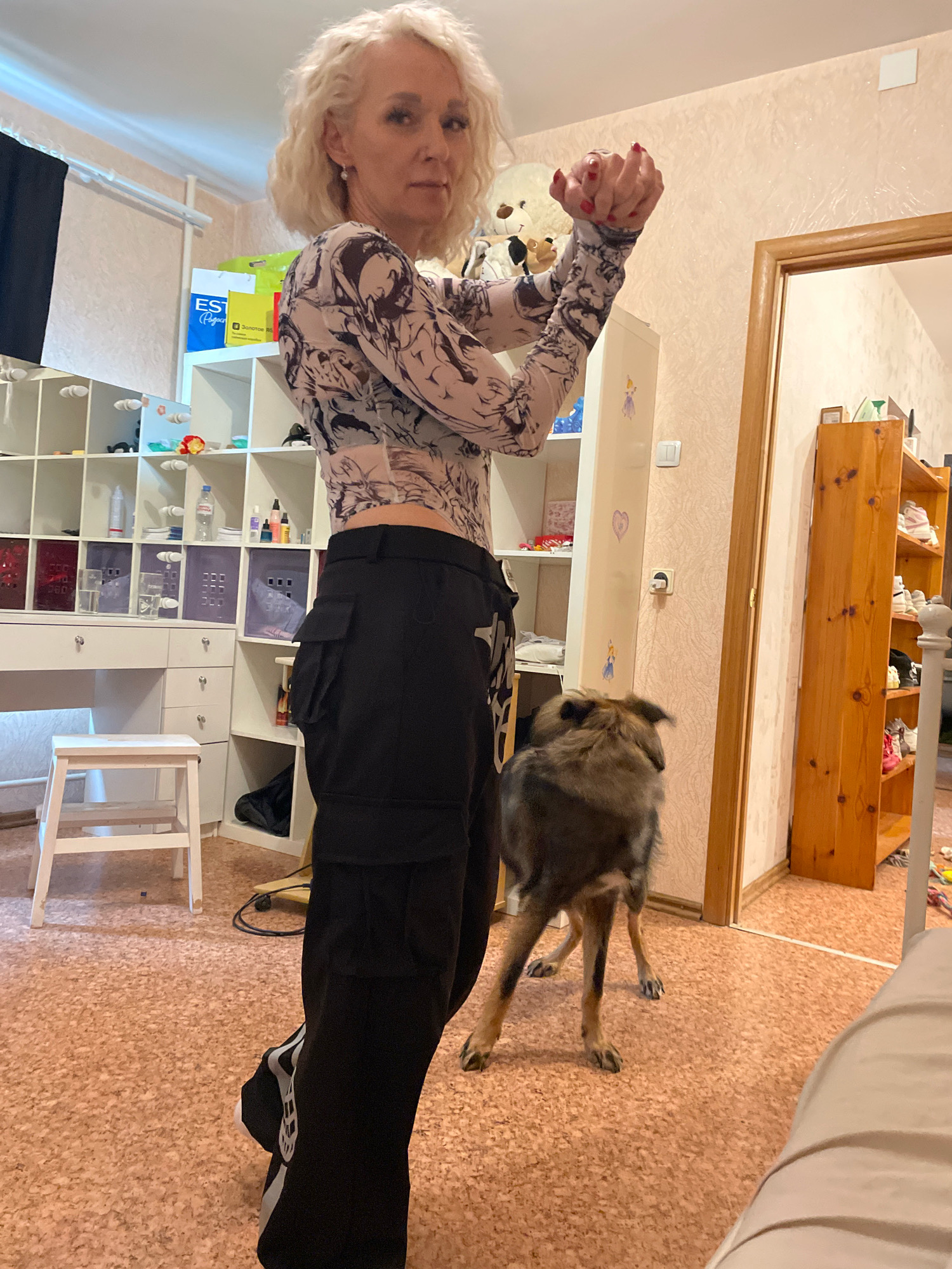 A woman with blonde curly hair stands indoors, facing the camera with her hands clasped in front of her chest. She is wearing a long-sleeved, patterned top and black cargo pants. To her right, a medium-sized dog with brown and gray fur stands looking away from the camera. The room appears to be a home with a large white shelving unit filled with various items, a white desk with a stool, and a wooden door leading to another room. The floor is covered in a light-colored cork material. The scene is set in Saint Petersburg, Russia.