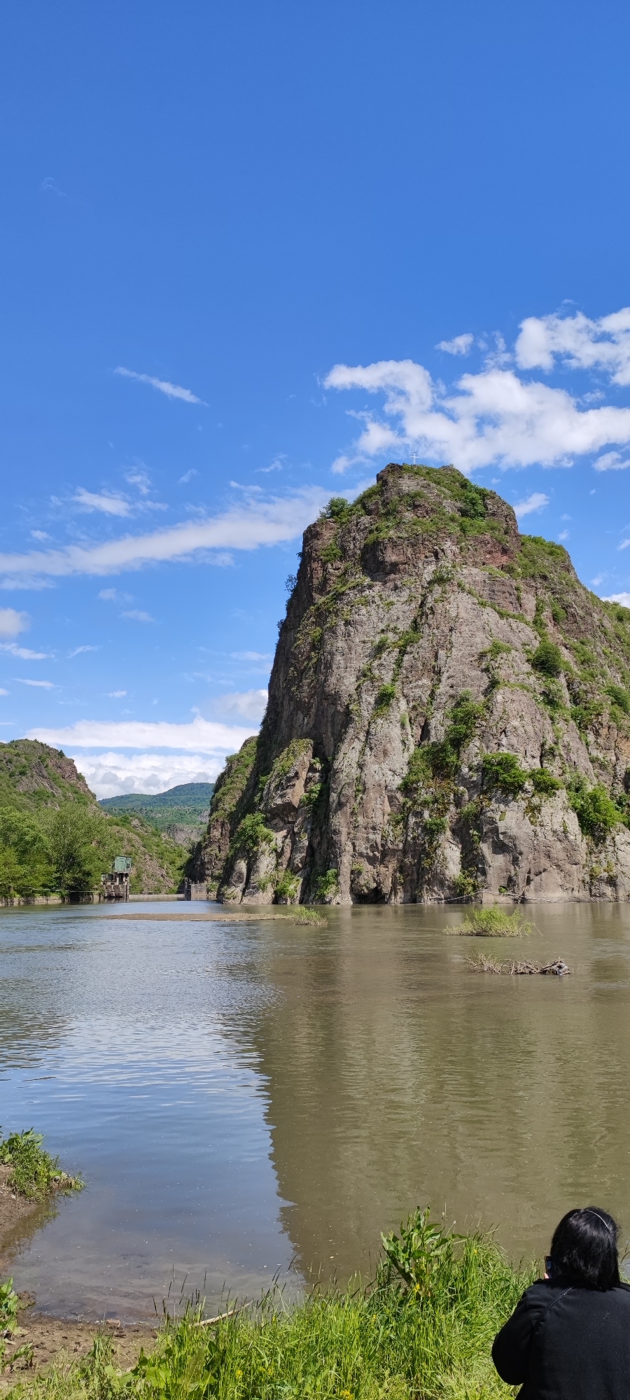 The image depicts a large, rocky mountain in the Armenian city of Arevatsag, partially submerged in a muddy river. The mountain has sparse vegetation and a cross at its peak. A small, building-like structure is visible along the river bank in the distance. The sky is blue with scattered clouds. In the foreground, a person is kneeling near the riverbank.