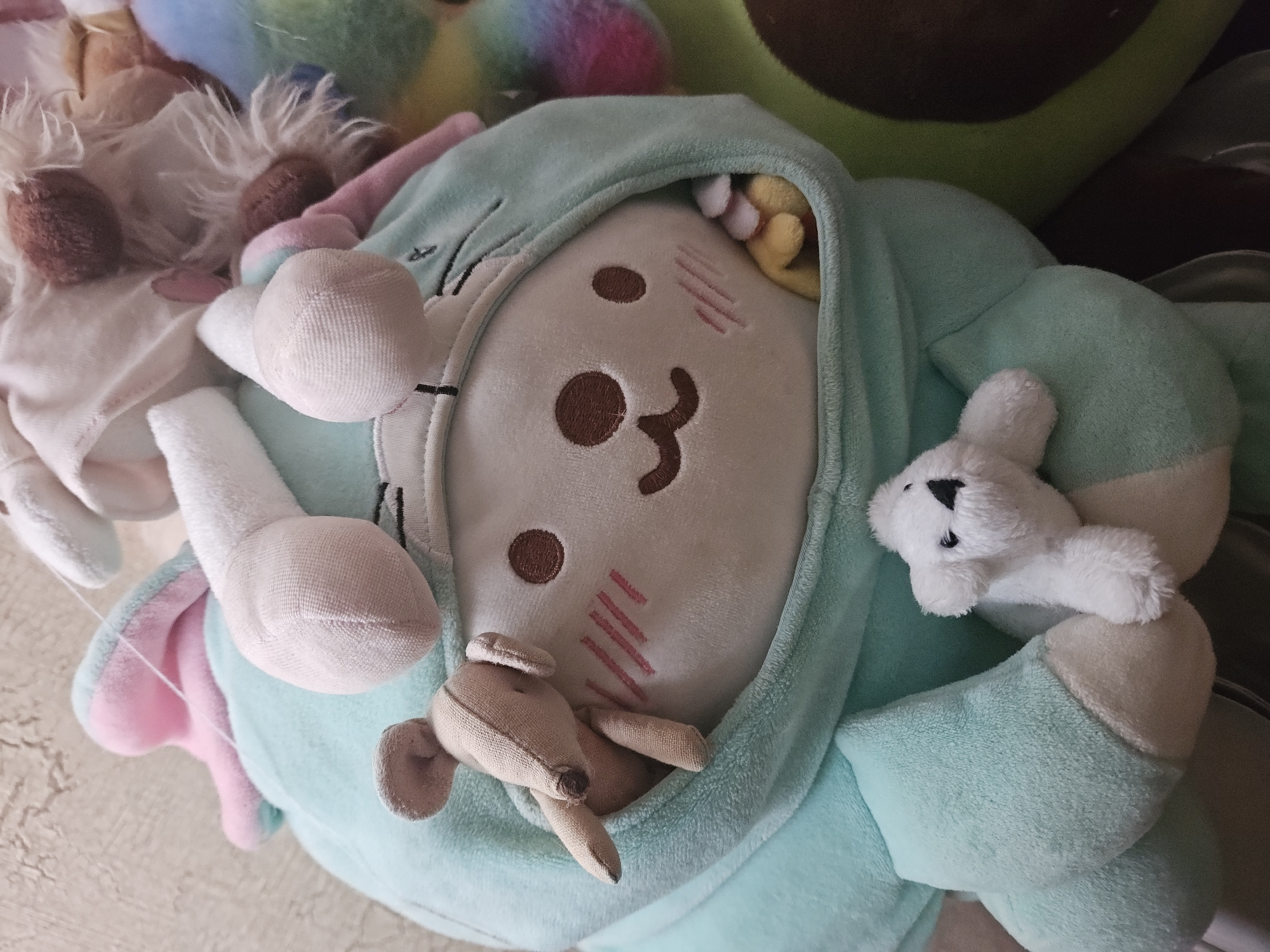 The image shows a close-up of several stuffed toys. A large, light blue plush toy with pink ears and a hood with white bunny ears is prominent. Inside the hood, a white plush face with brown eyes and a smiling mouth is visible, with pink blush marks on its cheeks. A small, tan stuffed mouse is peeking out from the side of the hood. Below the main plush toy, a small white polar bear stuffed toy is resting. In the background, a large green plush avocado toy is partially visible, along with other colorful stuffed toys. The toys are placed on a textured surface, possibly a wall or fabric. There is no information available about the location in the city of Ulyanovsk, Russia.