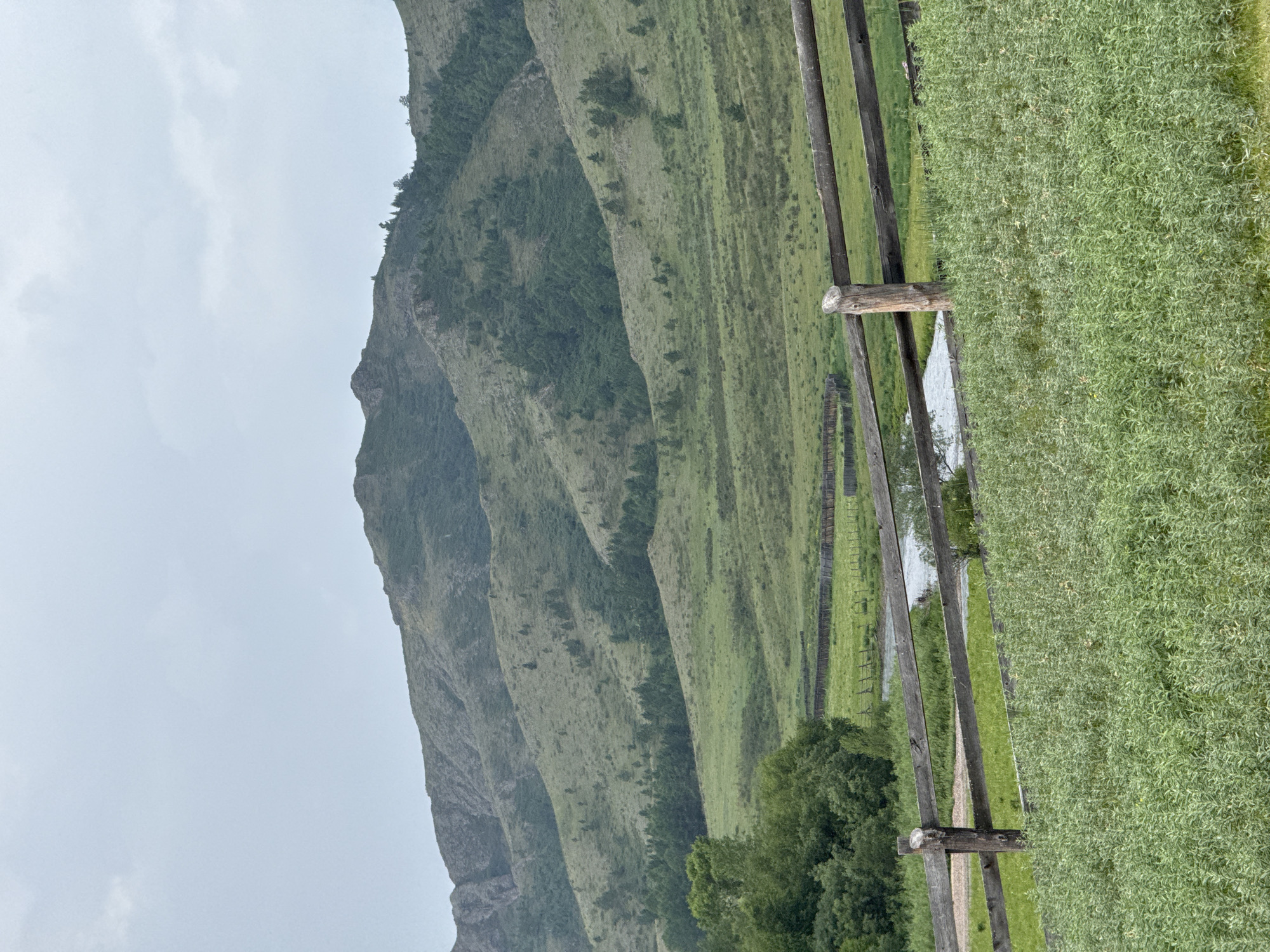 The image depicts a natural landscape under an overcast sky. In the foreground, a rustic wooden fence runs horizontally across the frame, constructed from rough-hewn logs. Beyond the fence, a body of water, likely a river or stream, meanders through a green meadow. Further in the distance, rolling hills covered in lush vegetation and scattered trees stretch towards a large mountain range in the background. The mountains are characterized by their rugged slopes, also dotted with trees. The overall scene suggests a rural or wilderness setting. The location is identified as Anchyl-Chon, Russia. There are no people or visible activity in the image.
